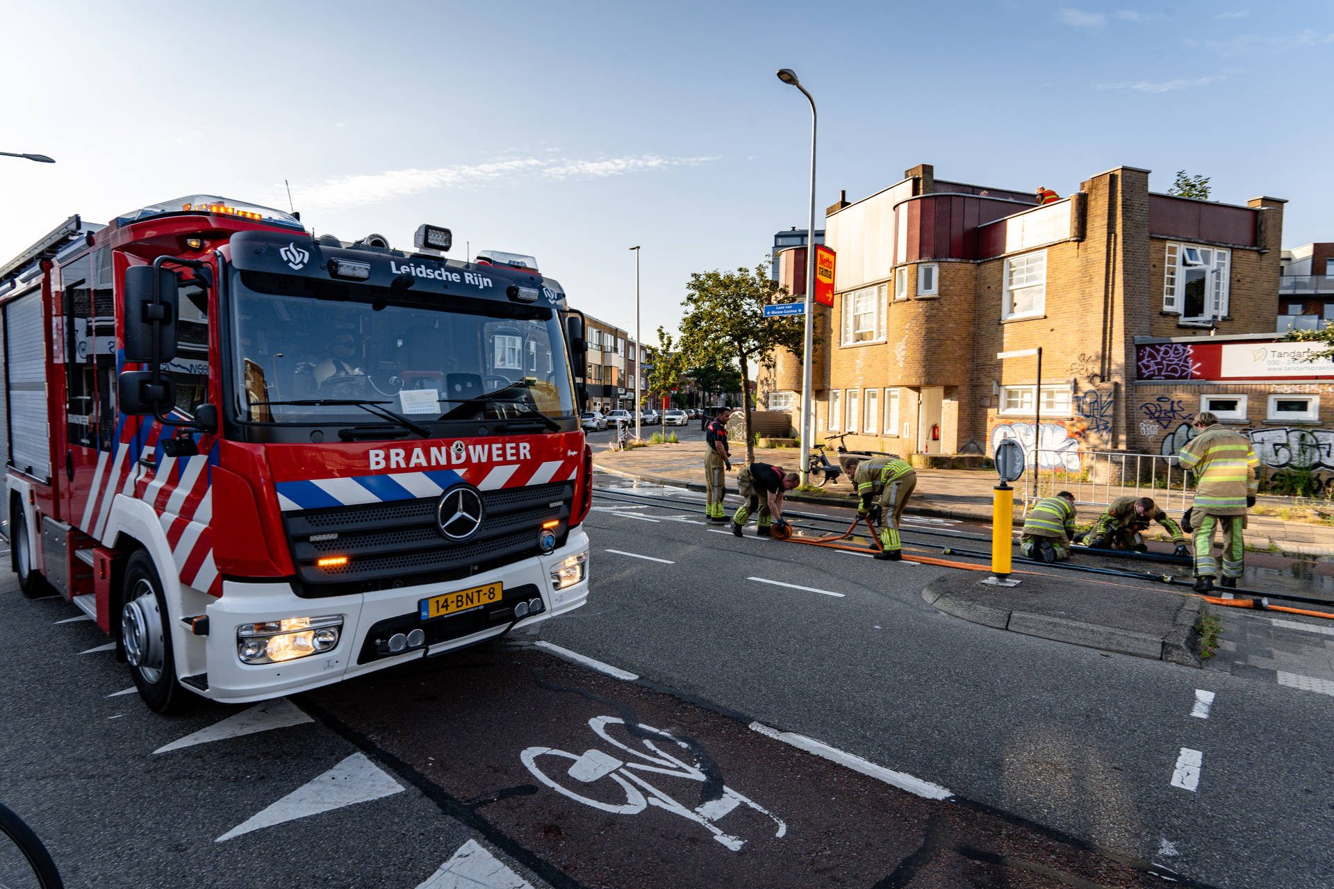 Firefighters prepare gear beside a large red fire truck on a sunny urban street at a crosswalk area.