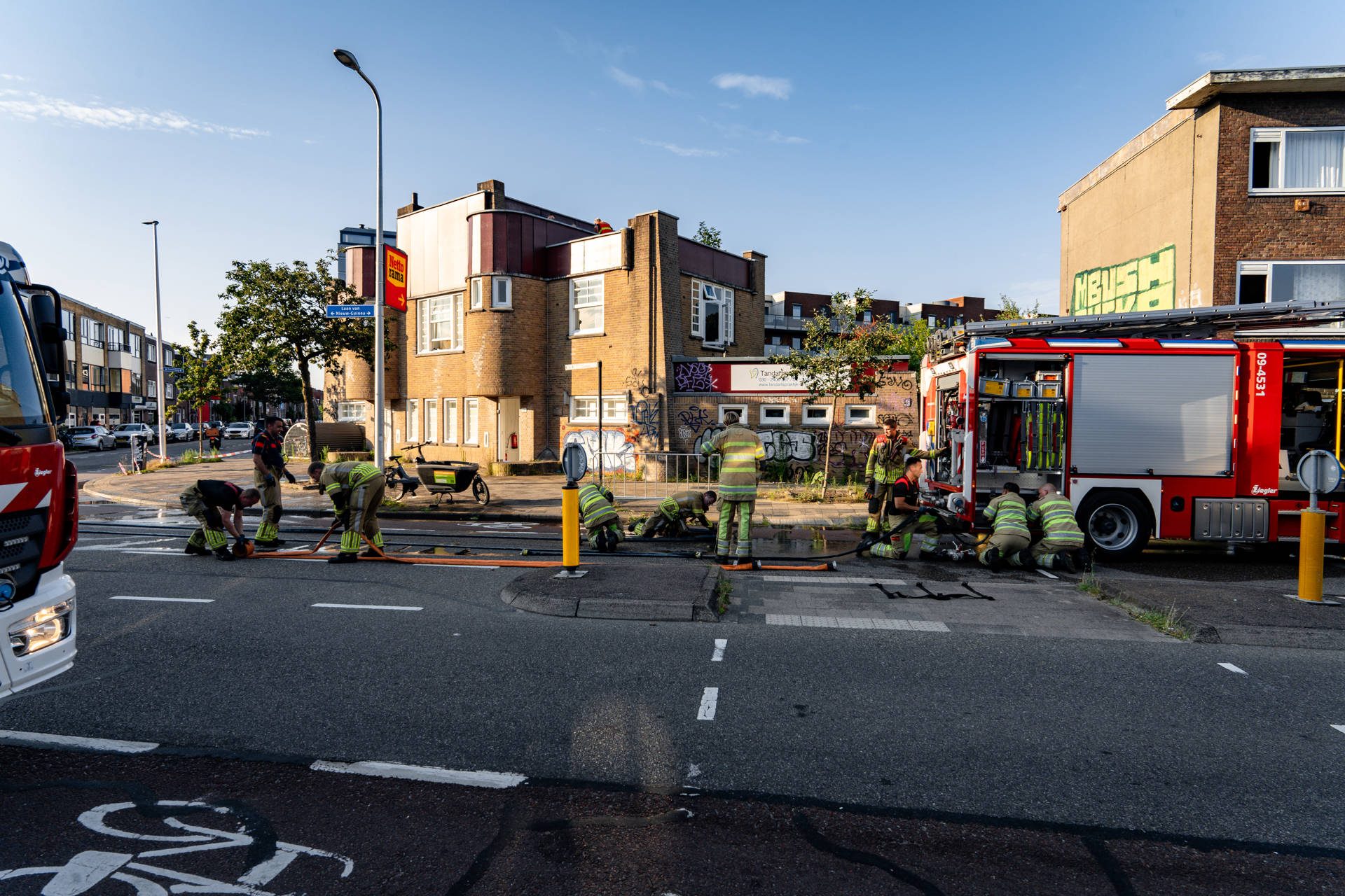 Firefighters in reflective gear lay hoses across a city street near a red fire engine, with a graffiti-covered building in the background.