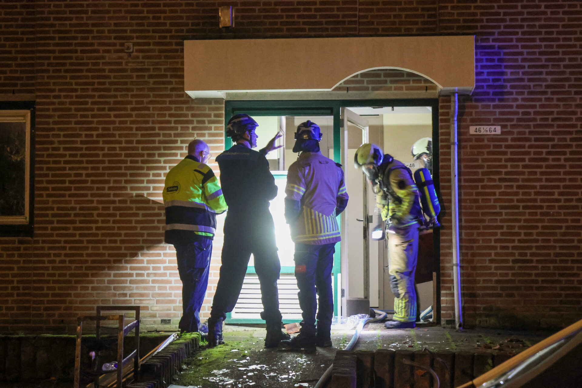 Emergency responders in reflective gear stand at a doorway in a brick building at night, hoses on the ground outside the entrance.