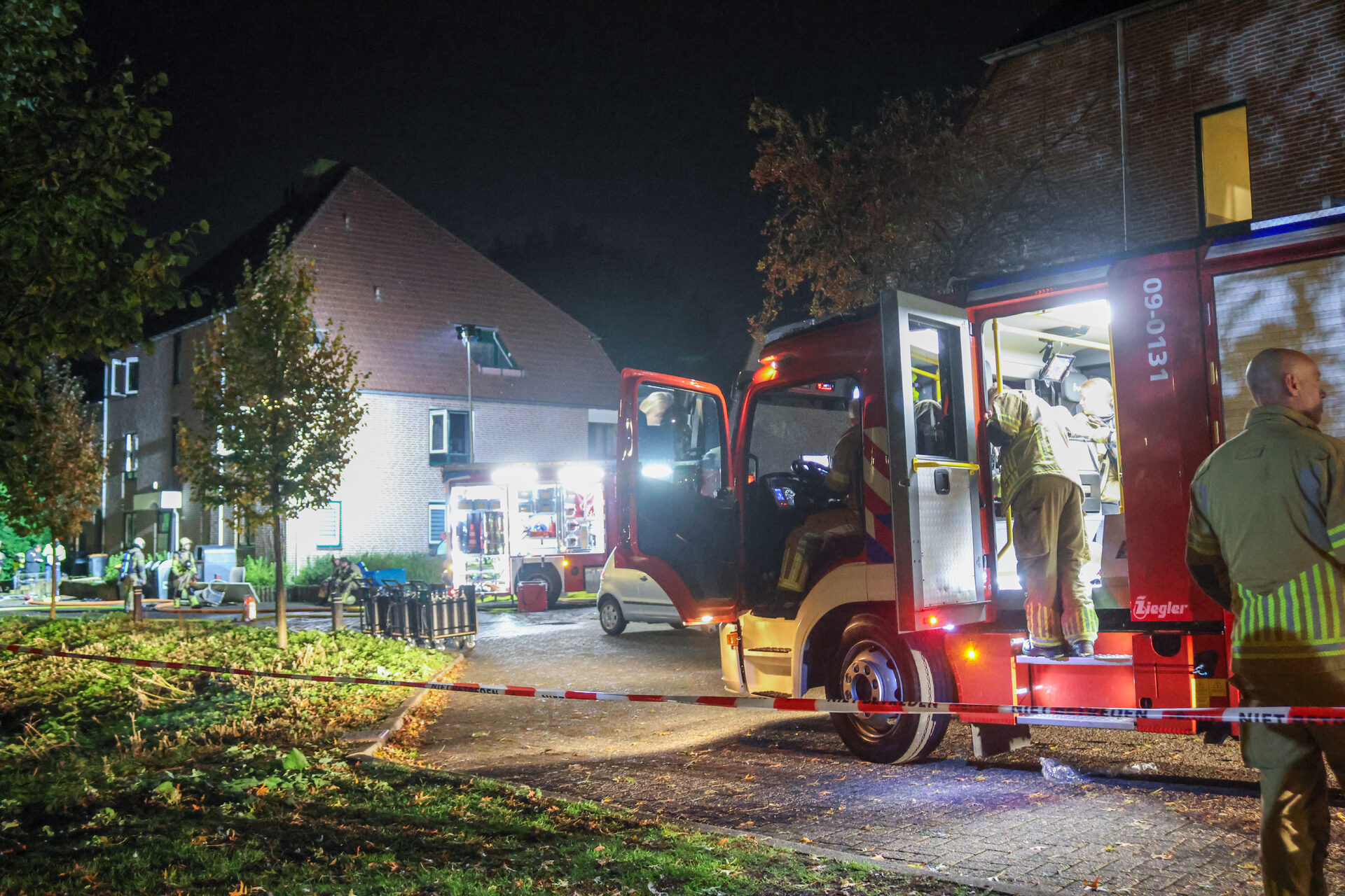 Firefighters and a red fire engine at a nighttime emergency scene outside a residential building, with caution tape and equipment on the ground.
