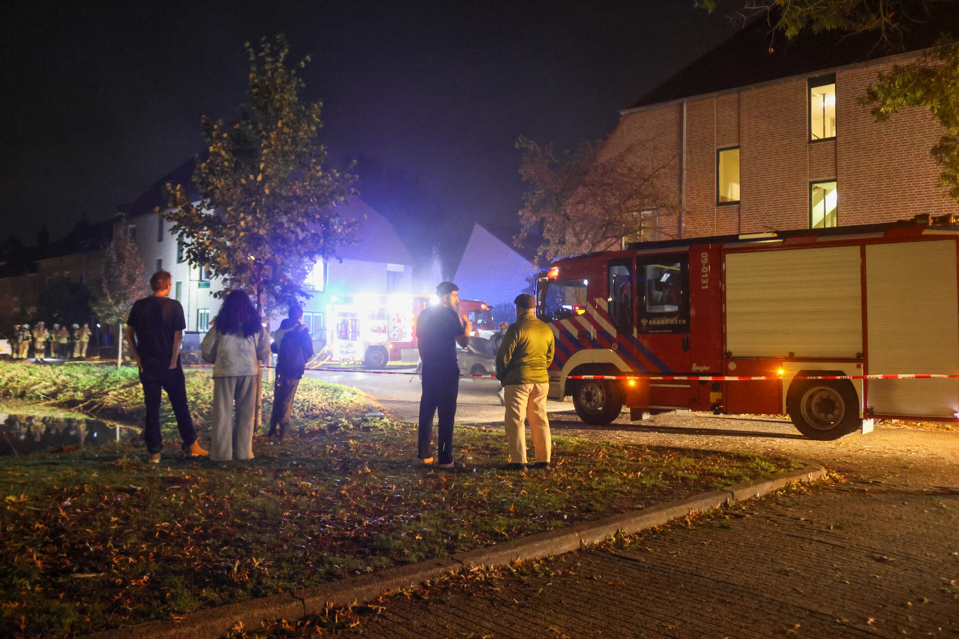 Night scene with a red fire truck and firefighters outside a brick apartment building; onlookers stand nearby as emergency lights flash.