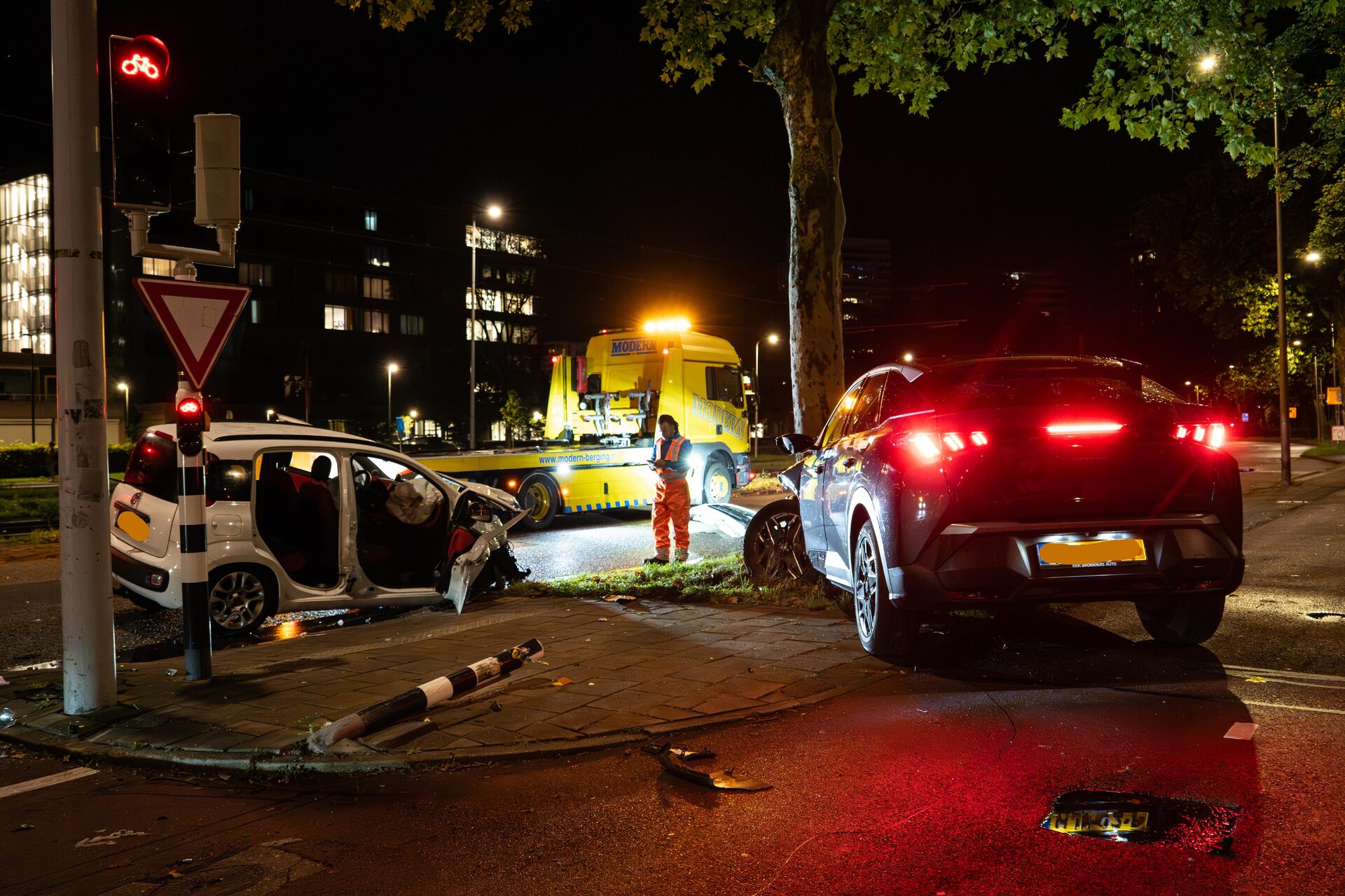 Nighttime road accident scene with a yellow tow truck and a worker in reflective gear assisting offboard vehicles beside a damaged white car and a red SUV. Debris on wet pavement is visible.