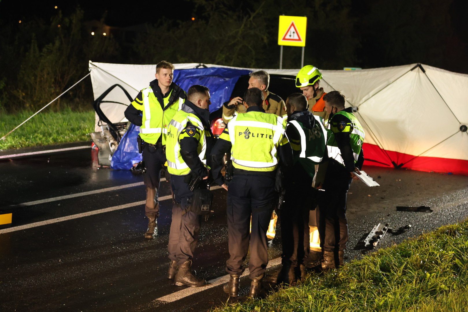 Night scene with police and firefighters in high-visibility jackets gathered around a white/blue tent on a wet road at night, assessing the scene.