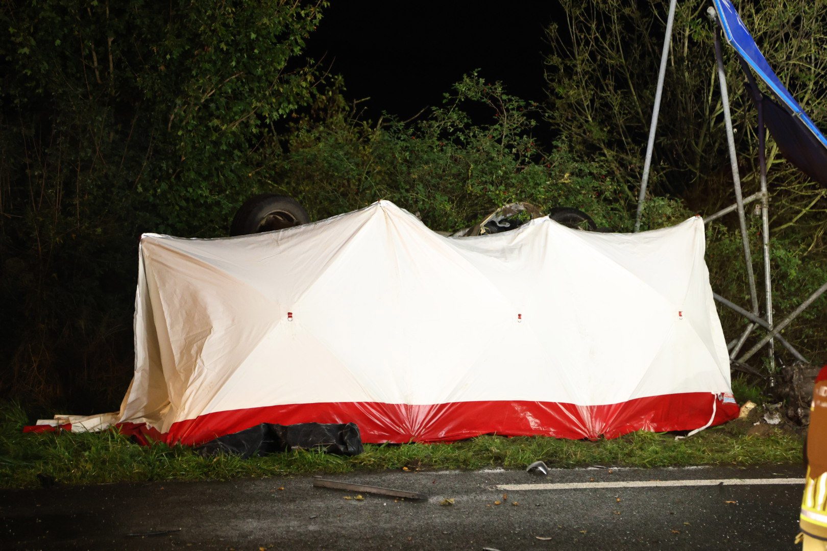 A large white tent with a red base lying on its side near a road at night, appears damaged or collapsed with scattered gear nearby.