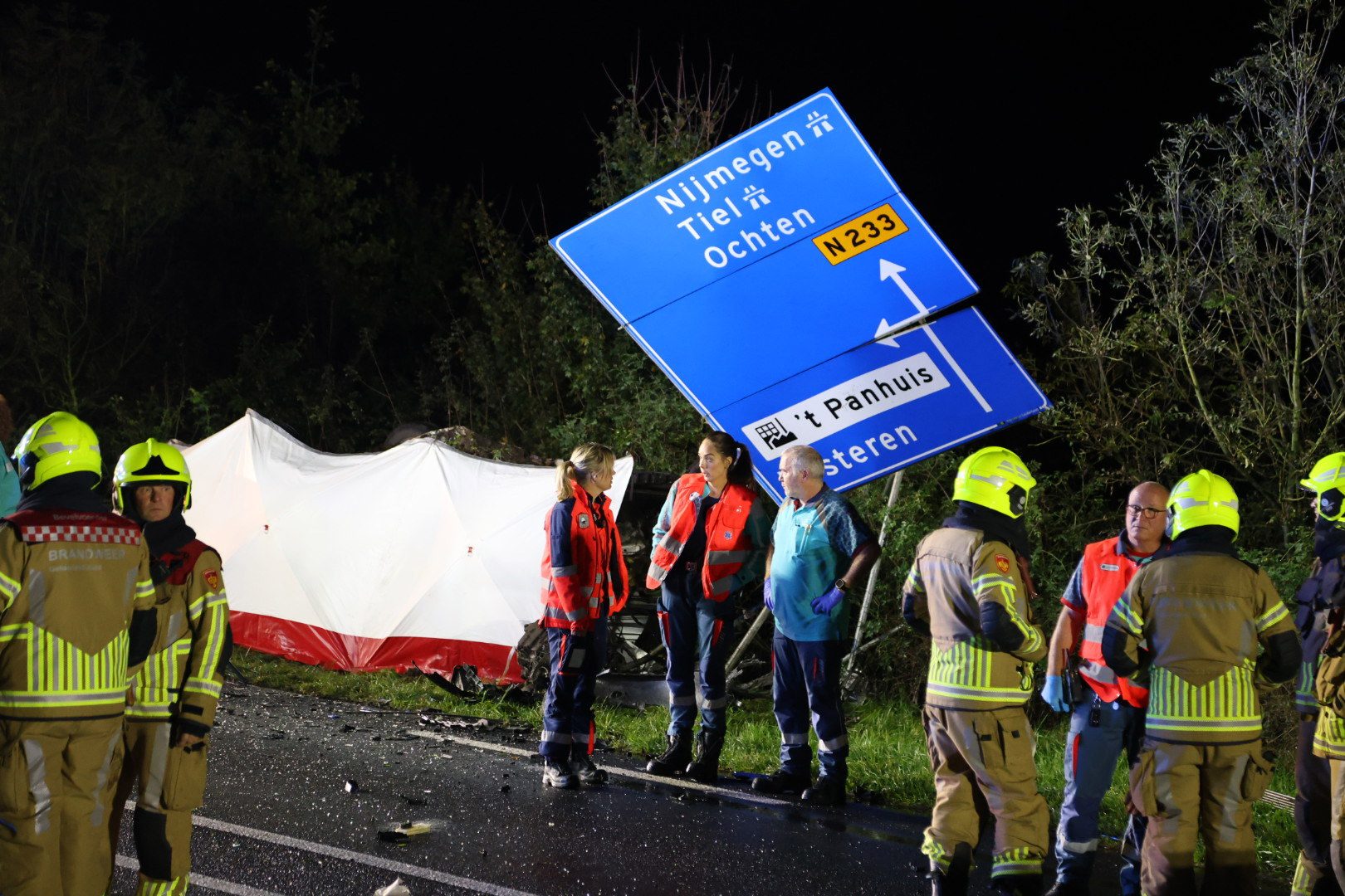 Emergency responders in high-visibility gear stand beside a tipped blue road sign on a dark highway at night, with a white-red tent behind them and scattered debris nearby.