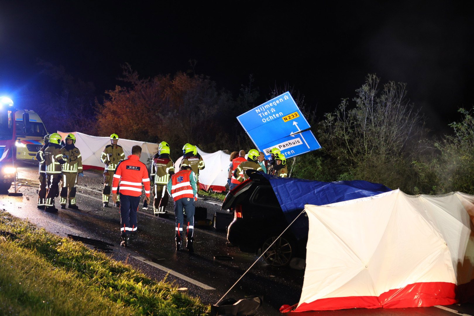 Nighttime accident scene with firefighters in yellow helmets and reflective gear surrounding a crashed car and red-and-white barriers/tents; blue emergency lights in the background.