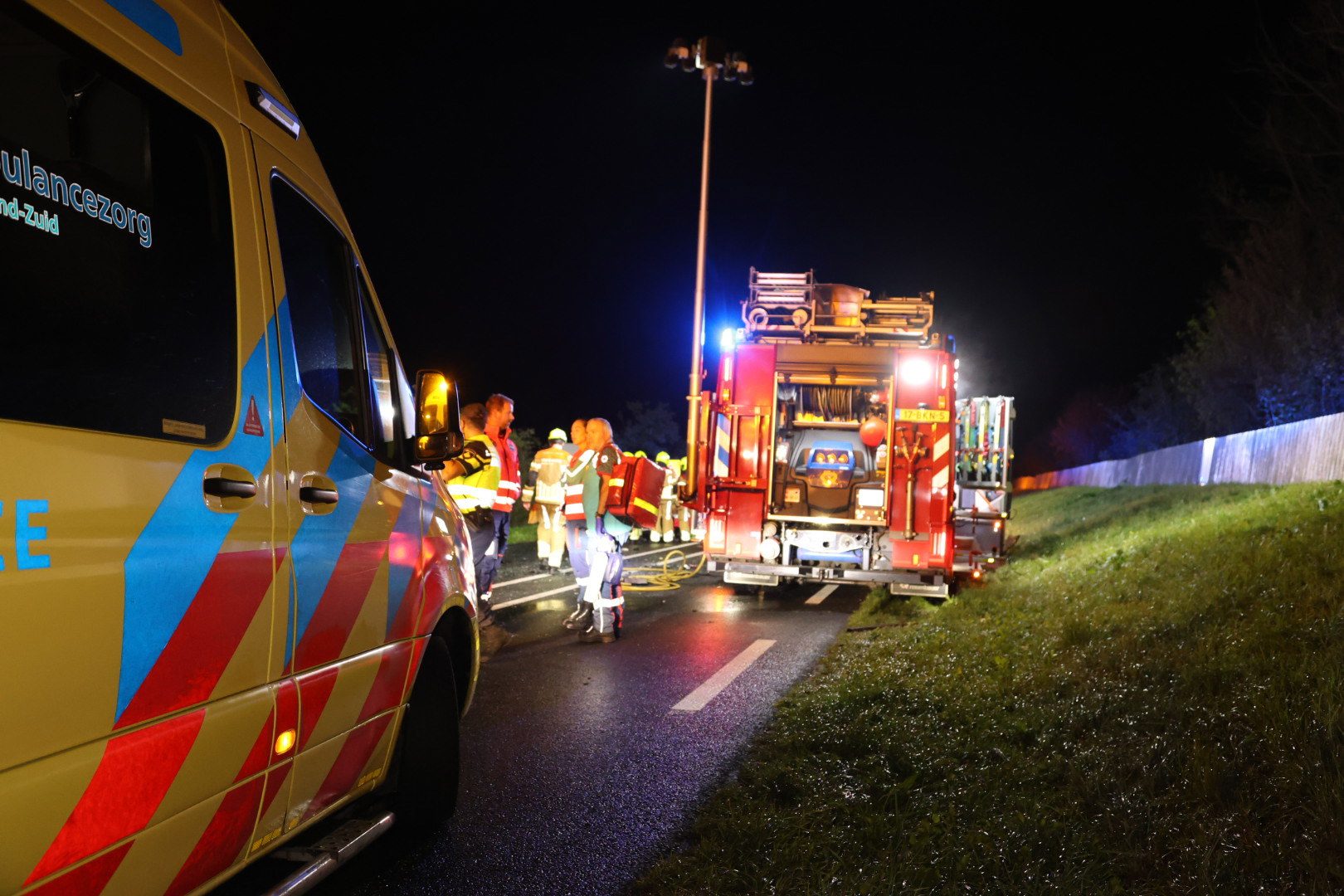 Nighttime emergency scene with an ambulance on the left and a red fire engine, responders in high-visibility gear gathered on a dark road.