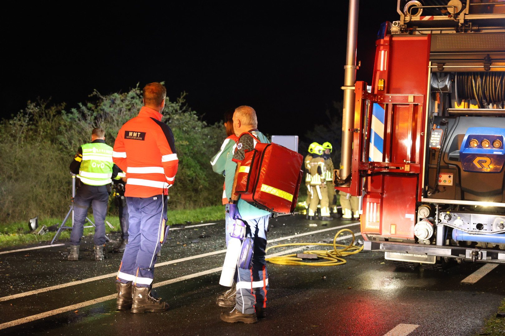 Emergency responders on a dark highway at night beside a red firetruck, with hoses and reflective gear visible.