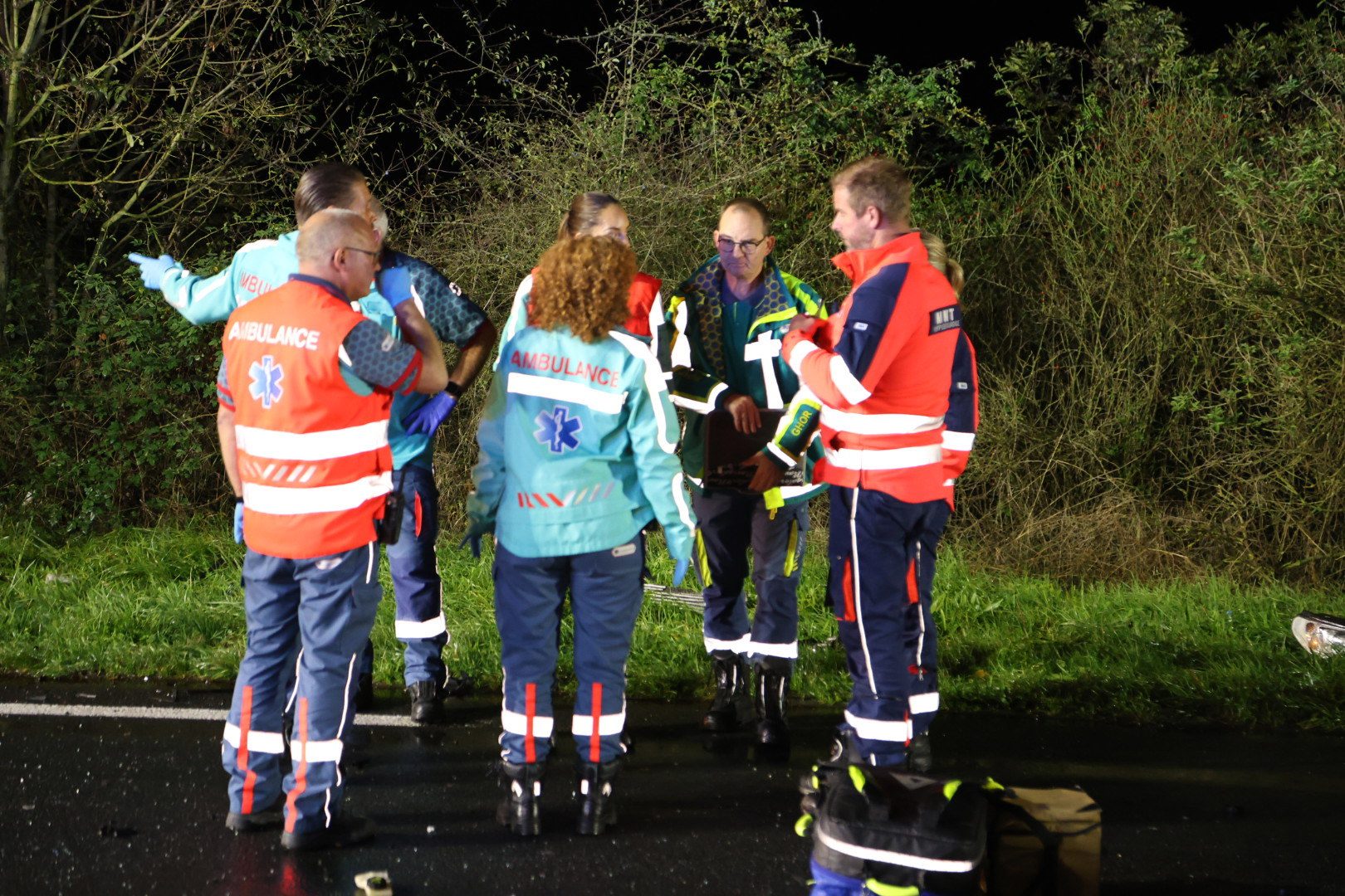 Paramedics in bright red and teal jackets stand in a circle at night, consulting around a patient on a stretcher beside a hedge.