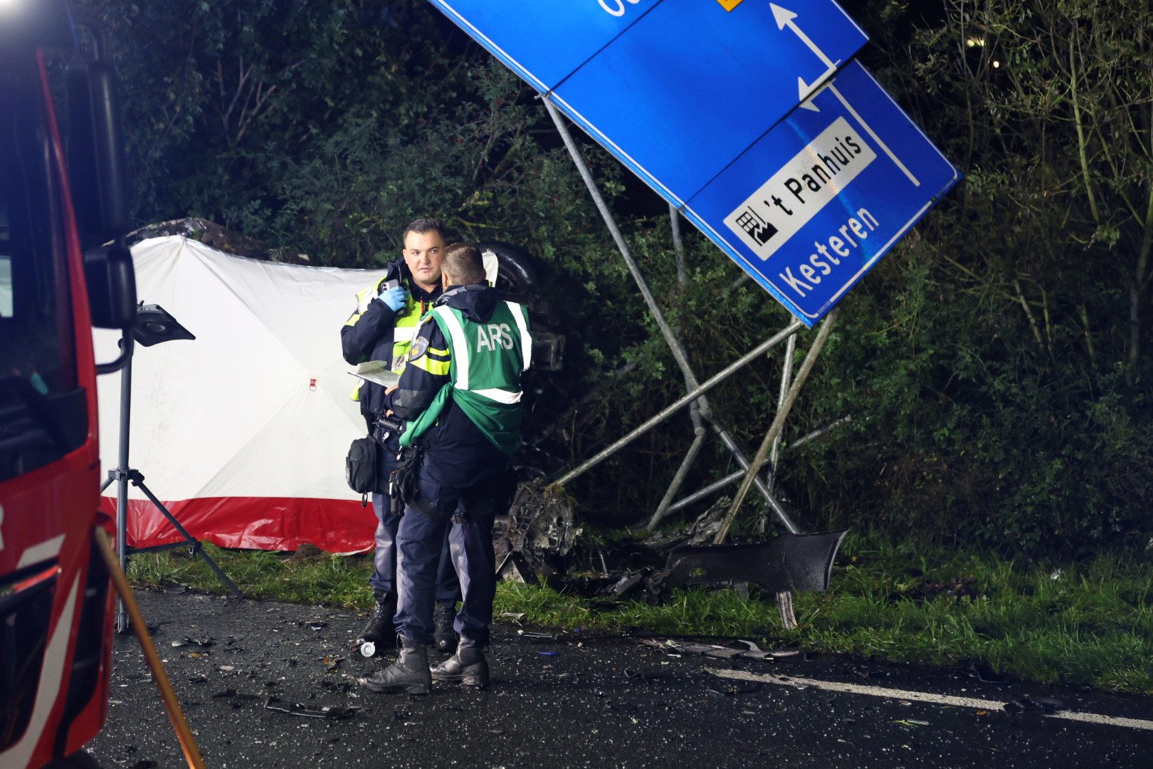 Two emergency responders in high‑visibility jackets talk beside a toppled blue road sign and a white tent at night.