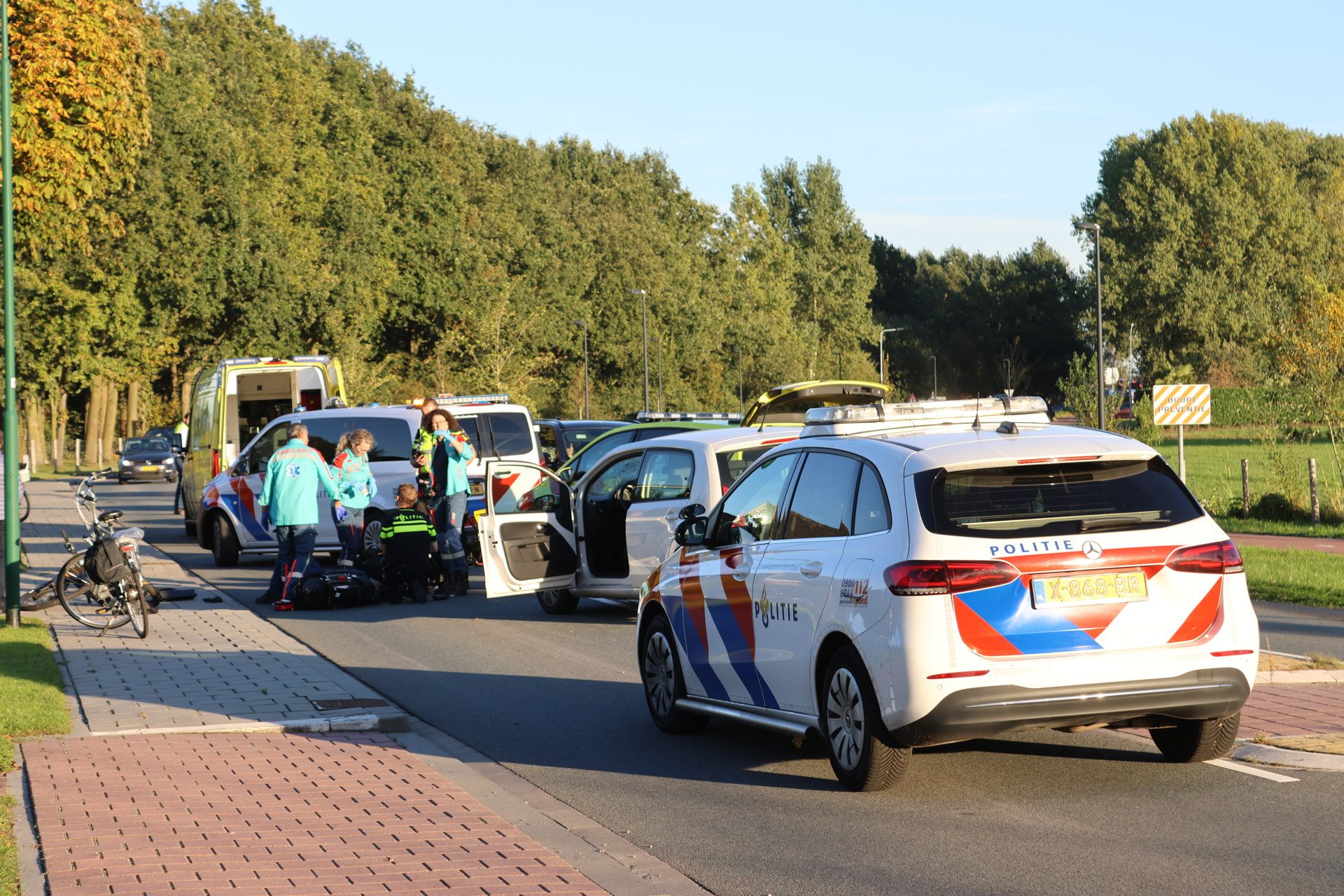 Police and medical responders assist at a roadside incident with multiple vehicles and bystanders on a sunny day.