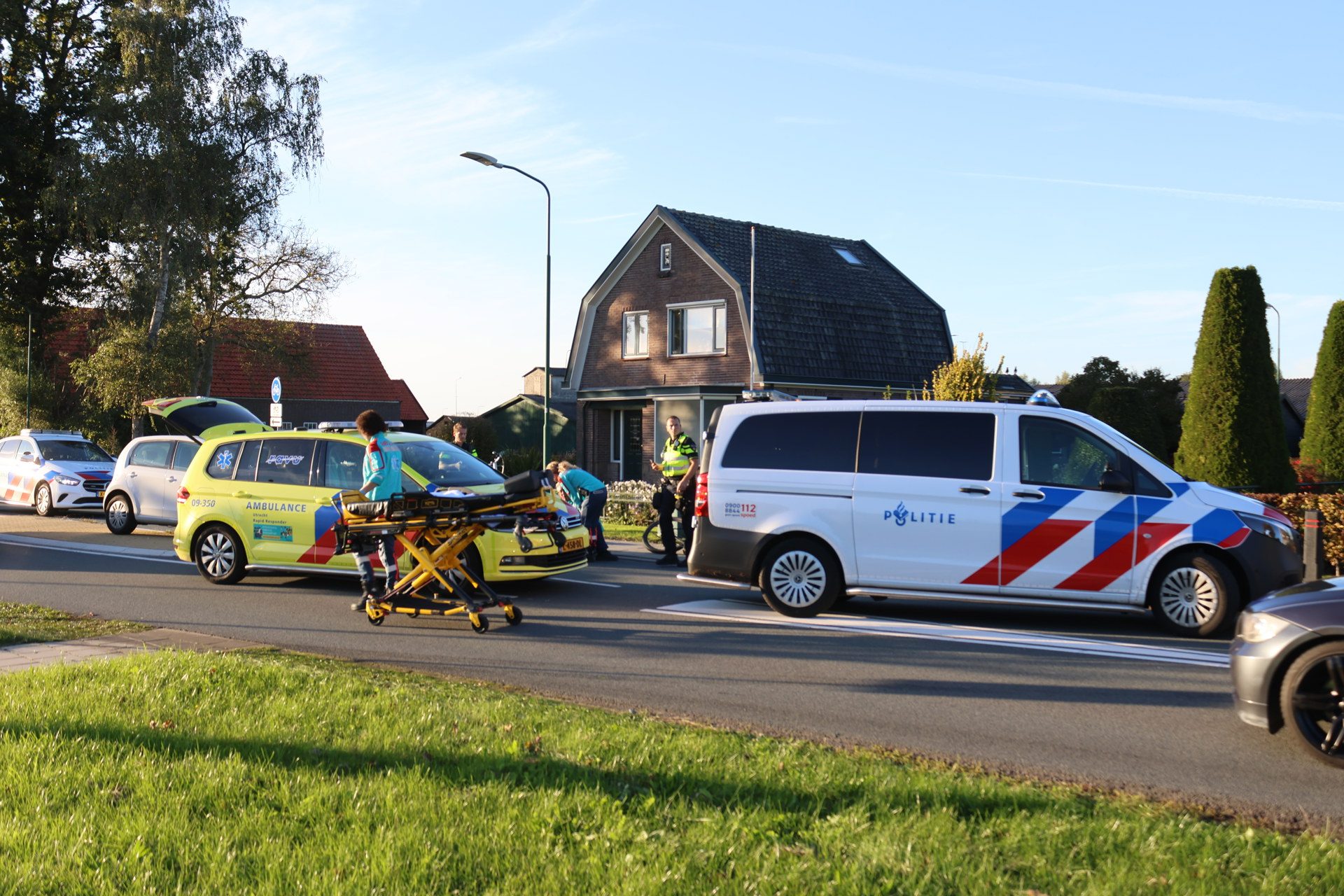 Emergency responders and a yellow ambulance with a stretcher on a wheeled cart in a residential street scene with a police van nearby.
