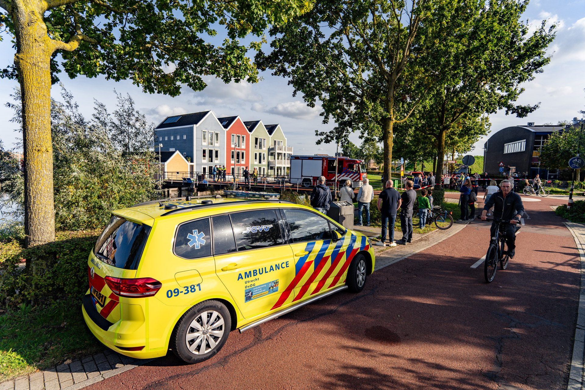 Yellow ambulance in the foreground on a red-brick street as bystanders gather near a canal and colorful row houses; a fire engine is visible in the background.