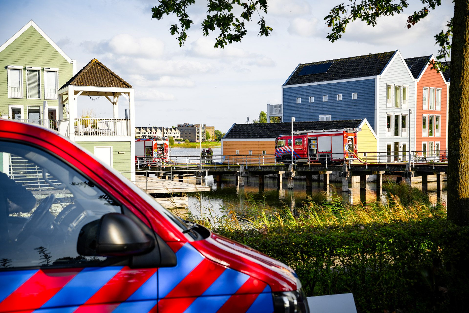 Emergency vehicles parked along a wooden bridge by a canal, with pastel waterfront houses in the background.