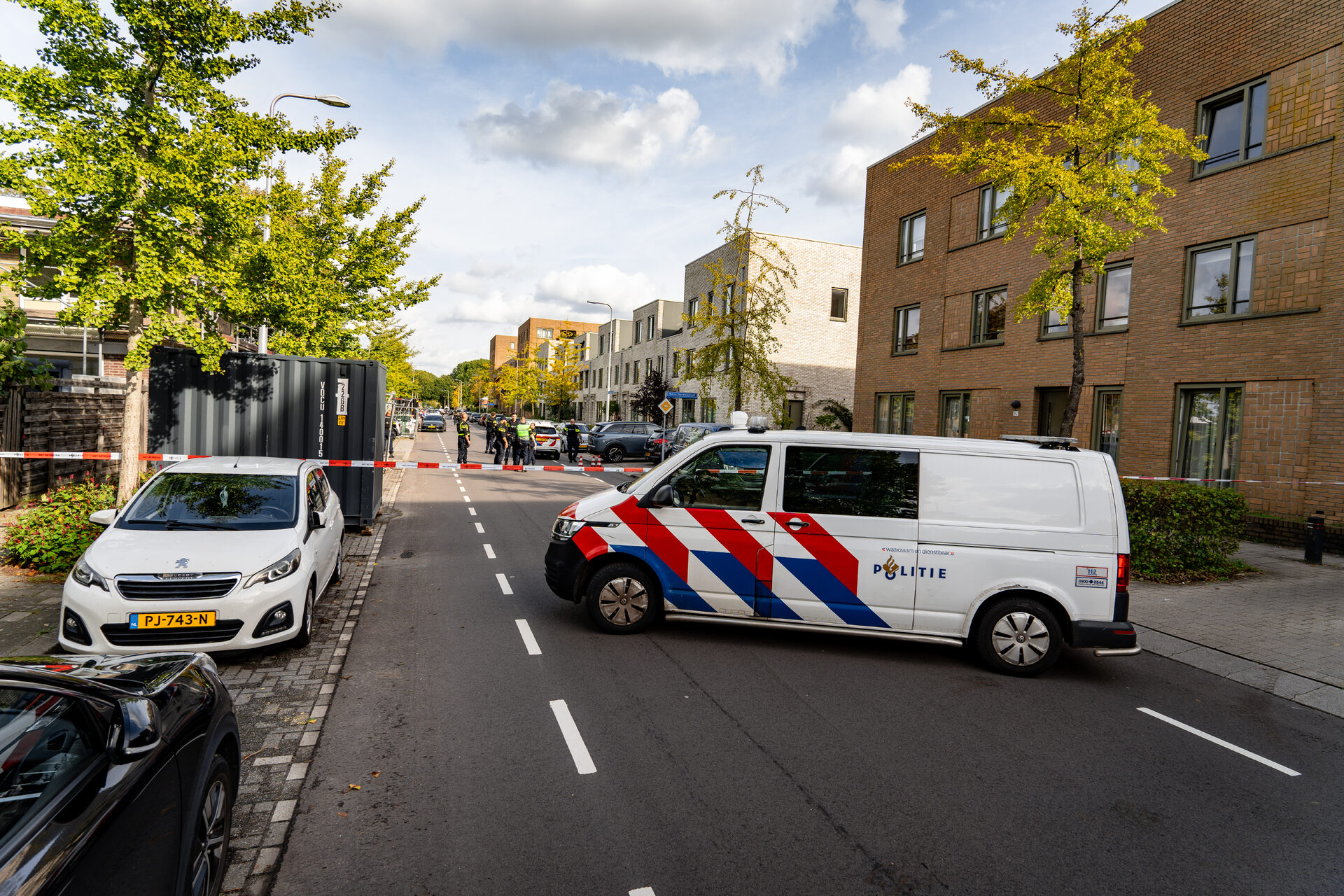 Police van blocks a city street with red-and-blue markings while officers stand behind red-and-white tape; scene cordoned off by responders on a sunny day in a residential area.