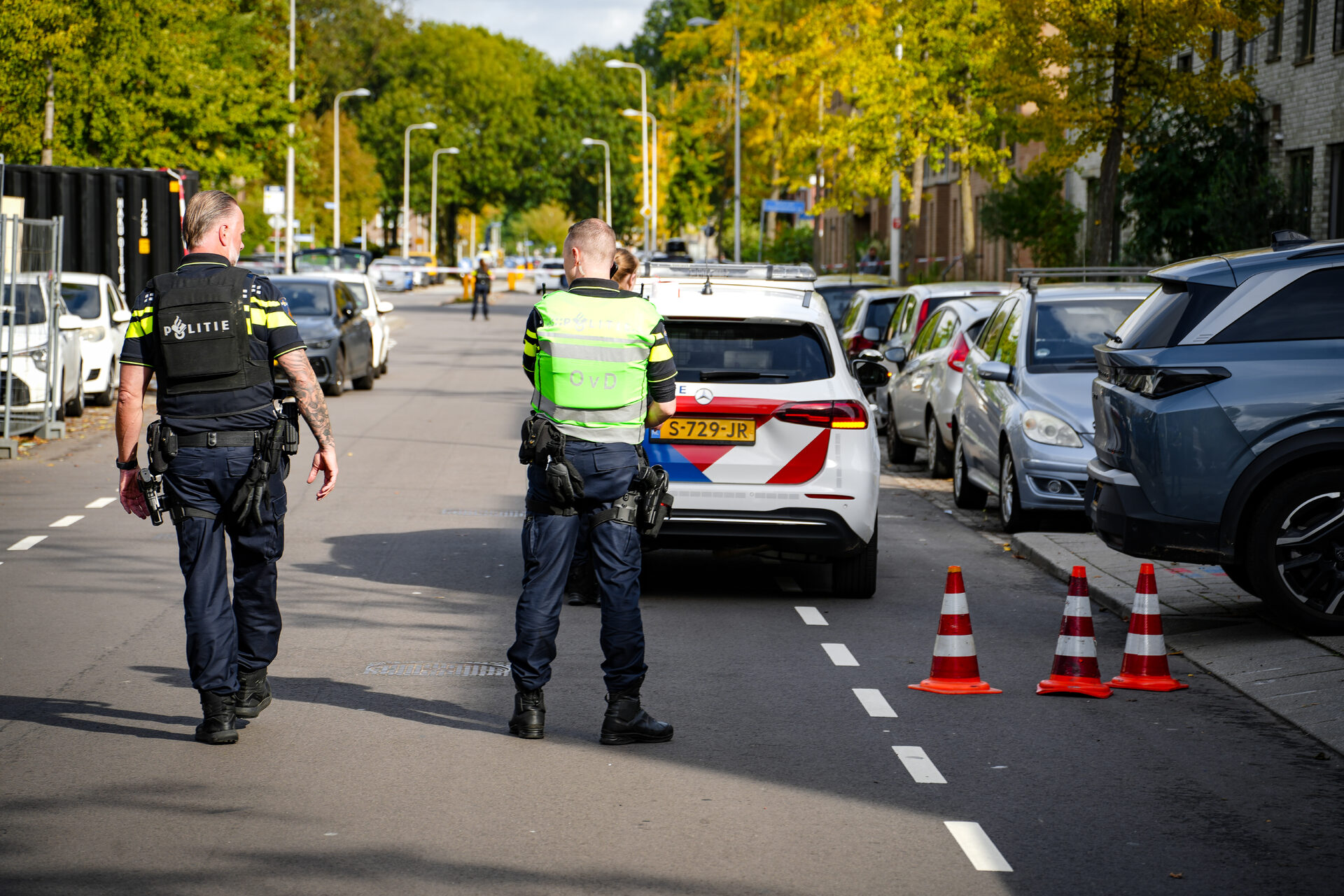 Two police officers stand in a cordoned-off city street, facing away, with a police car and traffic cones in view.