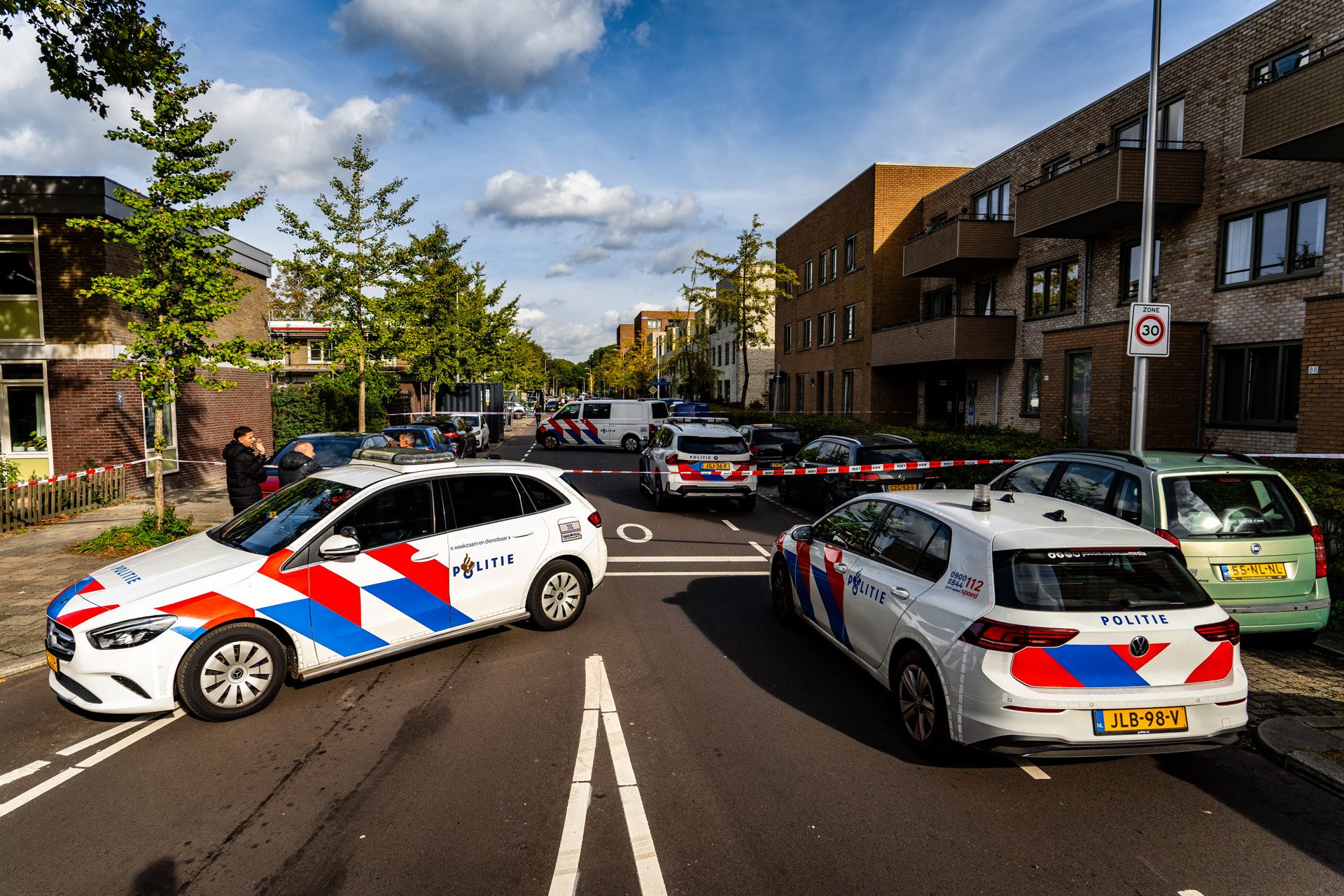 Politie cars block a residential street with police tape across the road and people talking nearby.