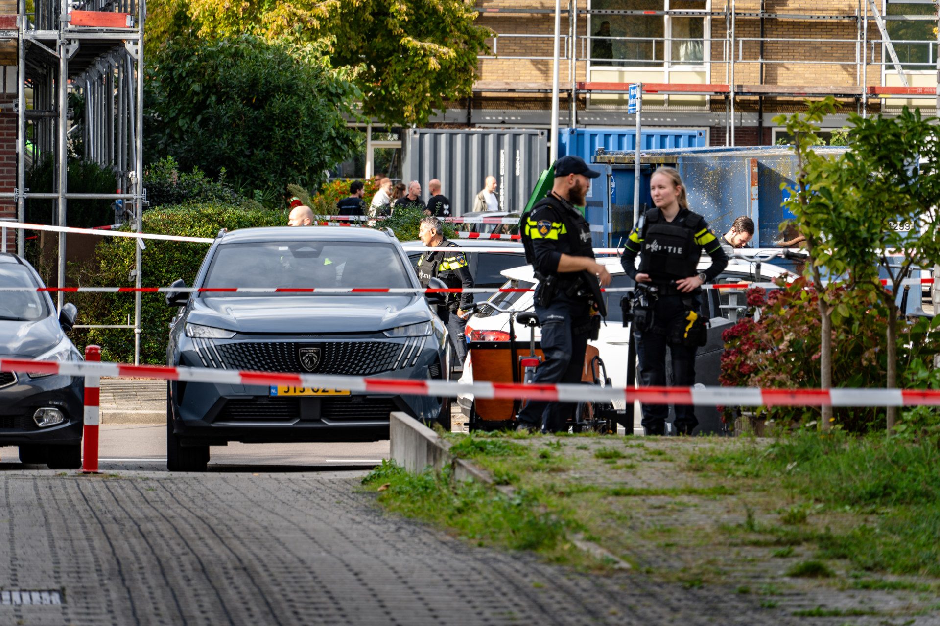 Police officers stand behind red-and-white barriers at a construction site with scaffolding and parked cars in the background.