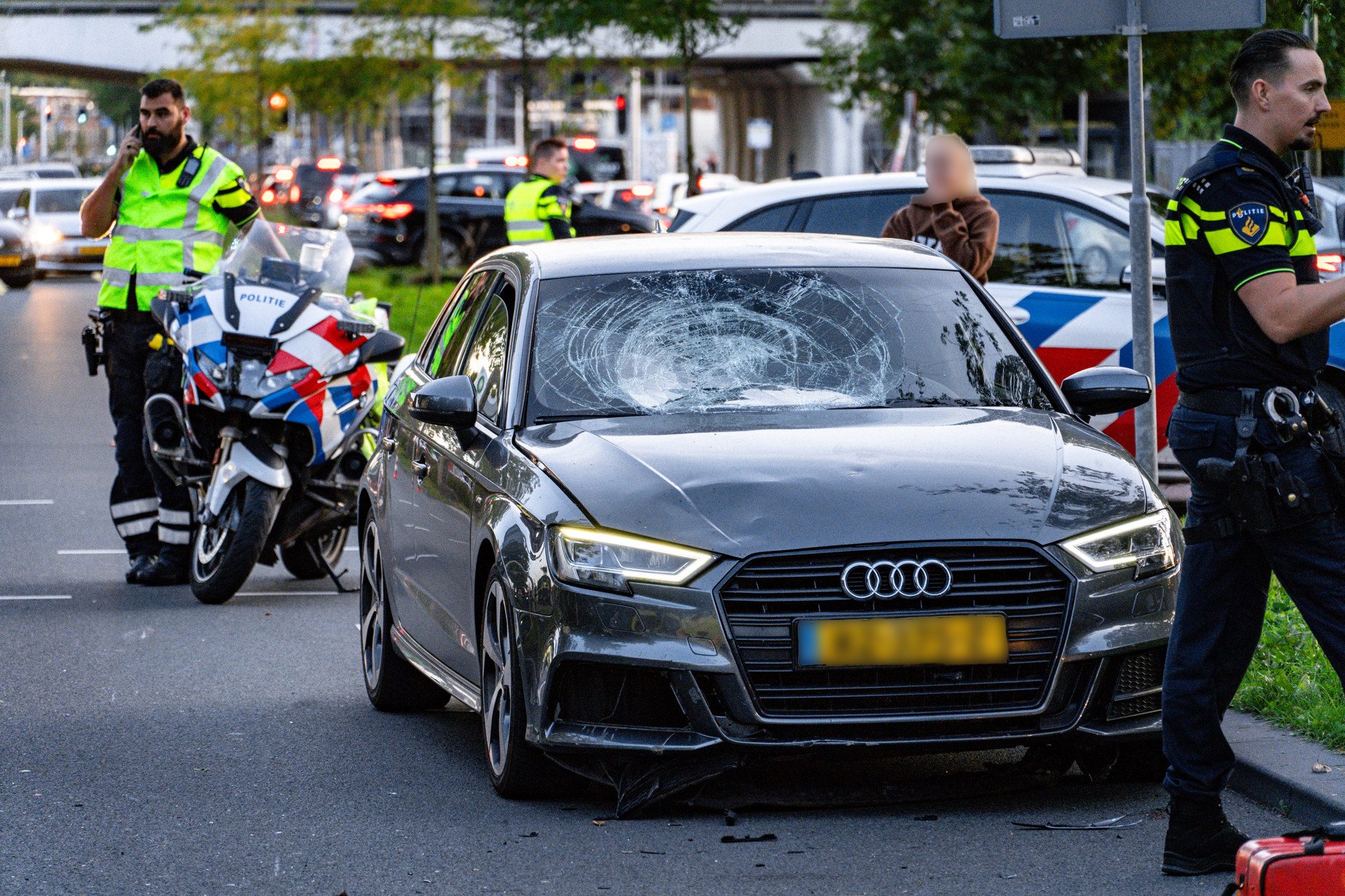 Grey Audi sedan with a large cracked windshield at a street checkpoint, police officers nearby and a police motorcycle present on the left.