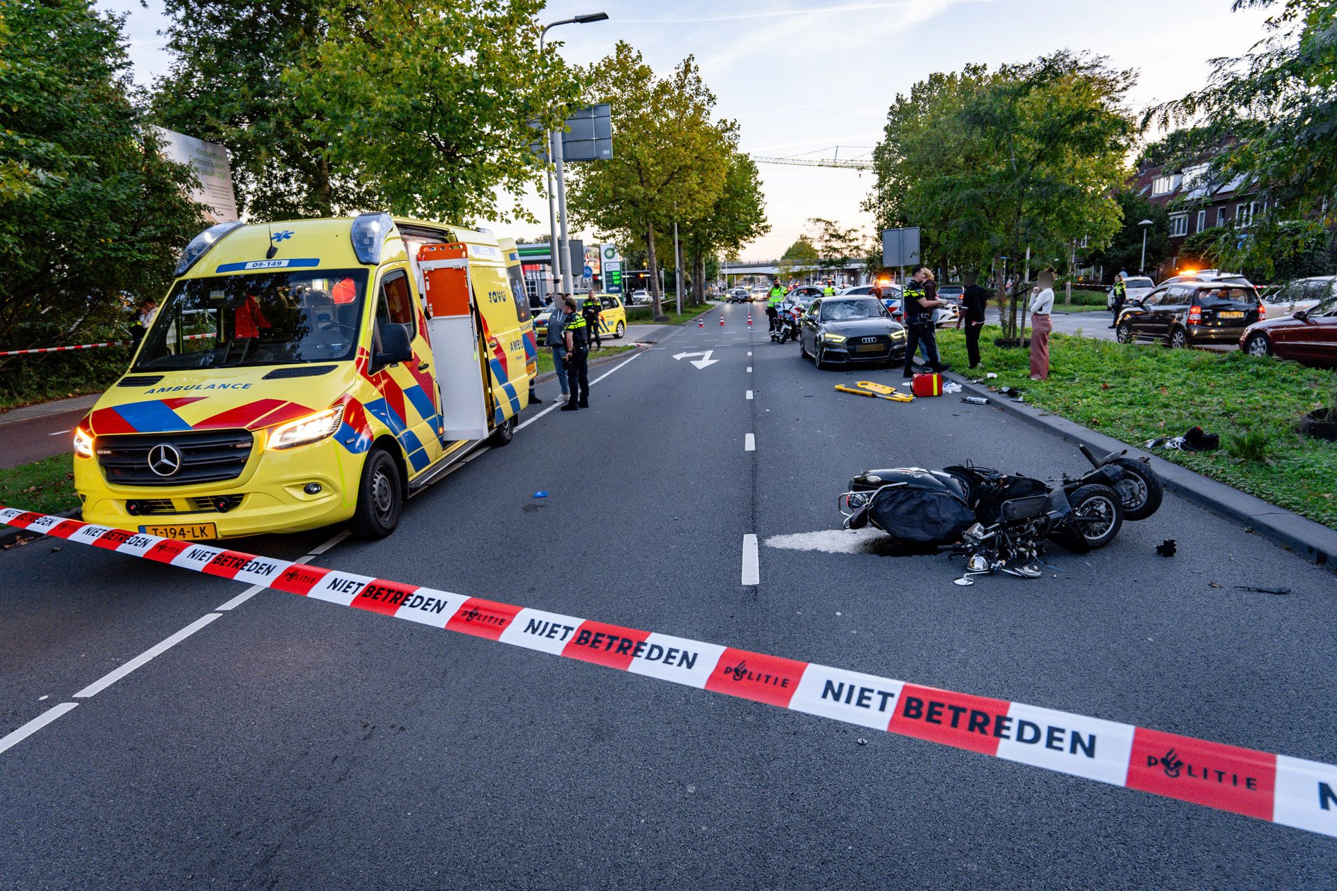 Emergency responders secure a weekday street after a motorcycle crash, with a yellow ambulance, police, and debris on the road and red-and-white tape reading 'NIET BETREDEN.'