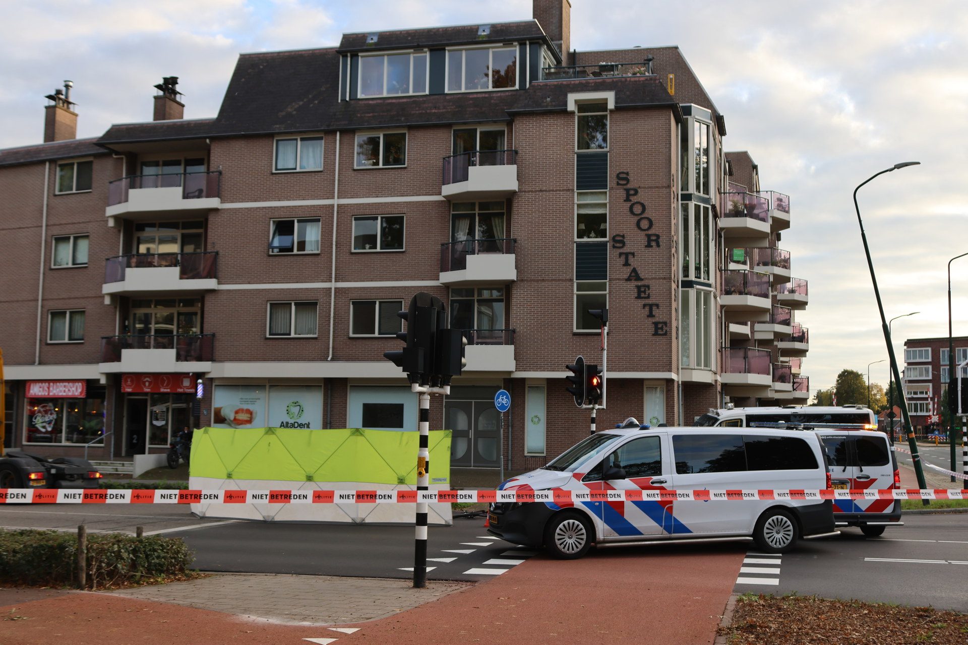 Multistory brick apartment building with balconies, a 'Spoorstate' sign on the side, and construction barriers in the street; emergency vehicles present nearby.