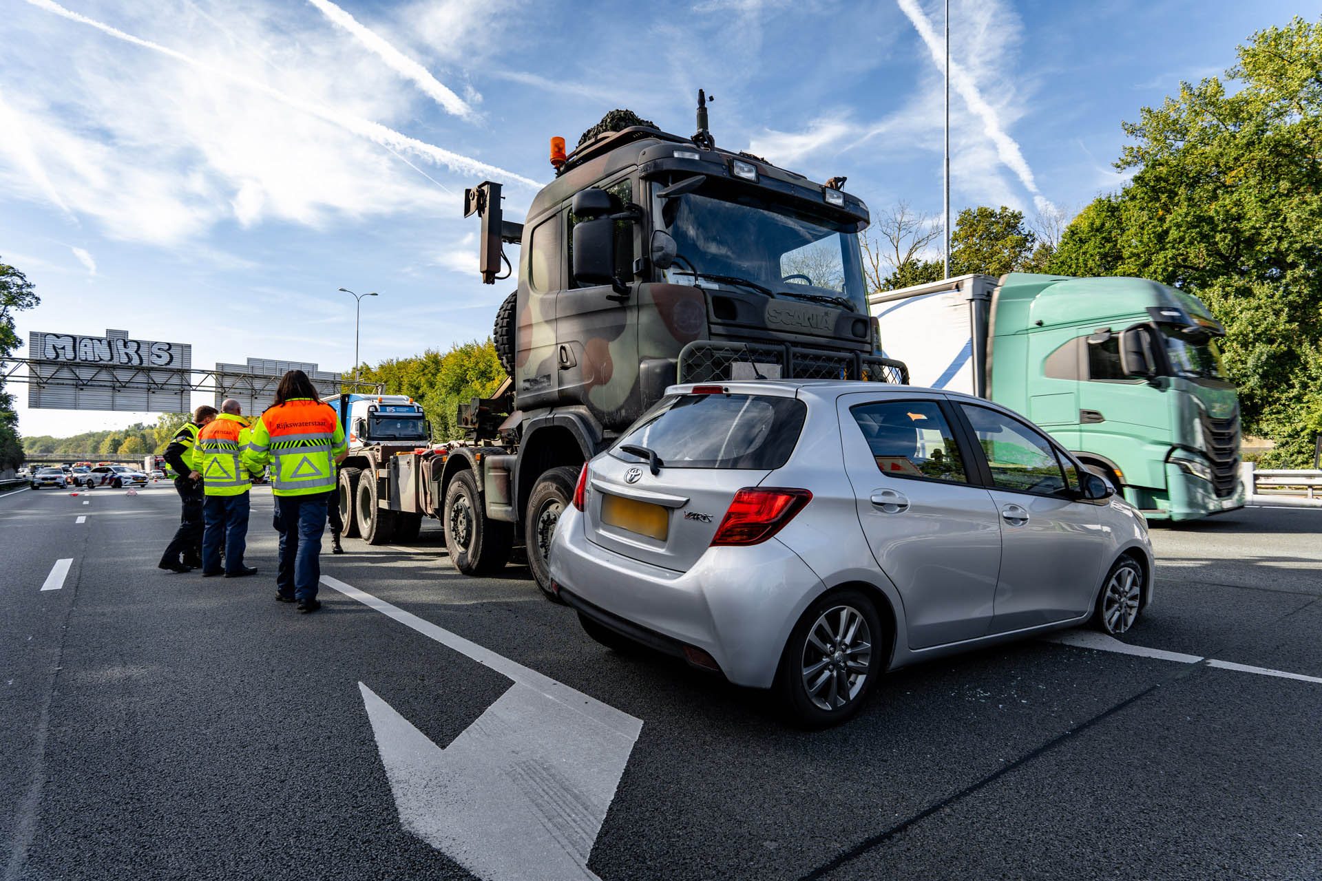 Highway incident with a camouflaged heavy truck on a trailer and workers in high-visibility jackets assessing the scene.