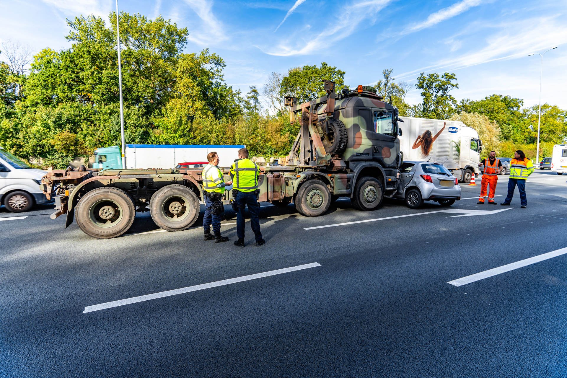 Highway scene with a camouflaged military-style truck on a trailer; two workers in high-visibility jackets converse nearby as traffic passes.