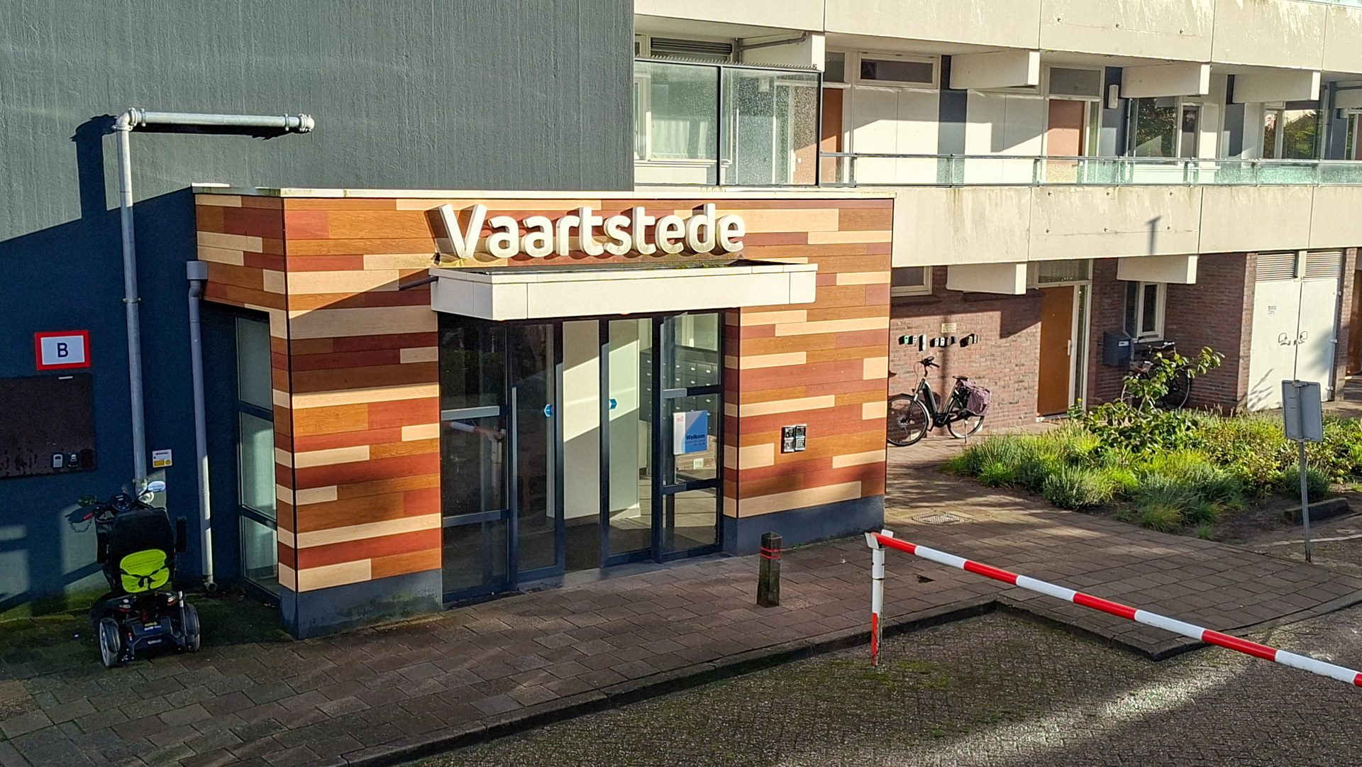 Entrance to Vaartstede building with a striped wooden façade, glass doors, and a bicycle parked nearby near a barrier gate.