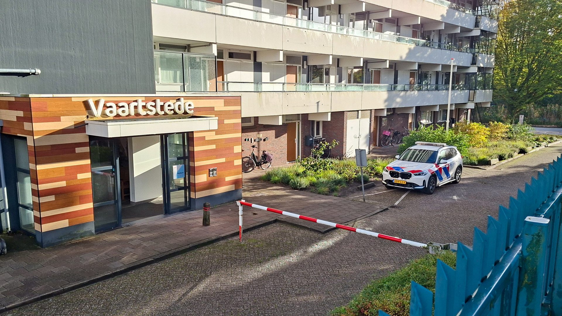Front of a residential building with a 'Vaartstede' sign, bikes near the entrance, and a police car in the parking area.