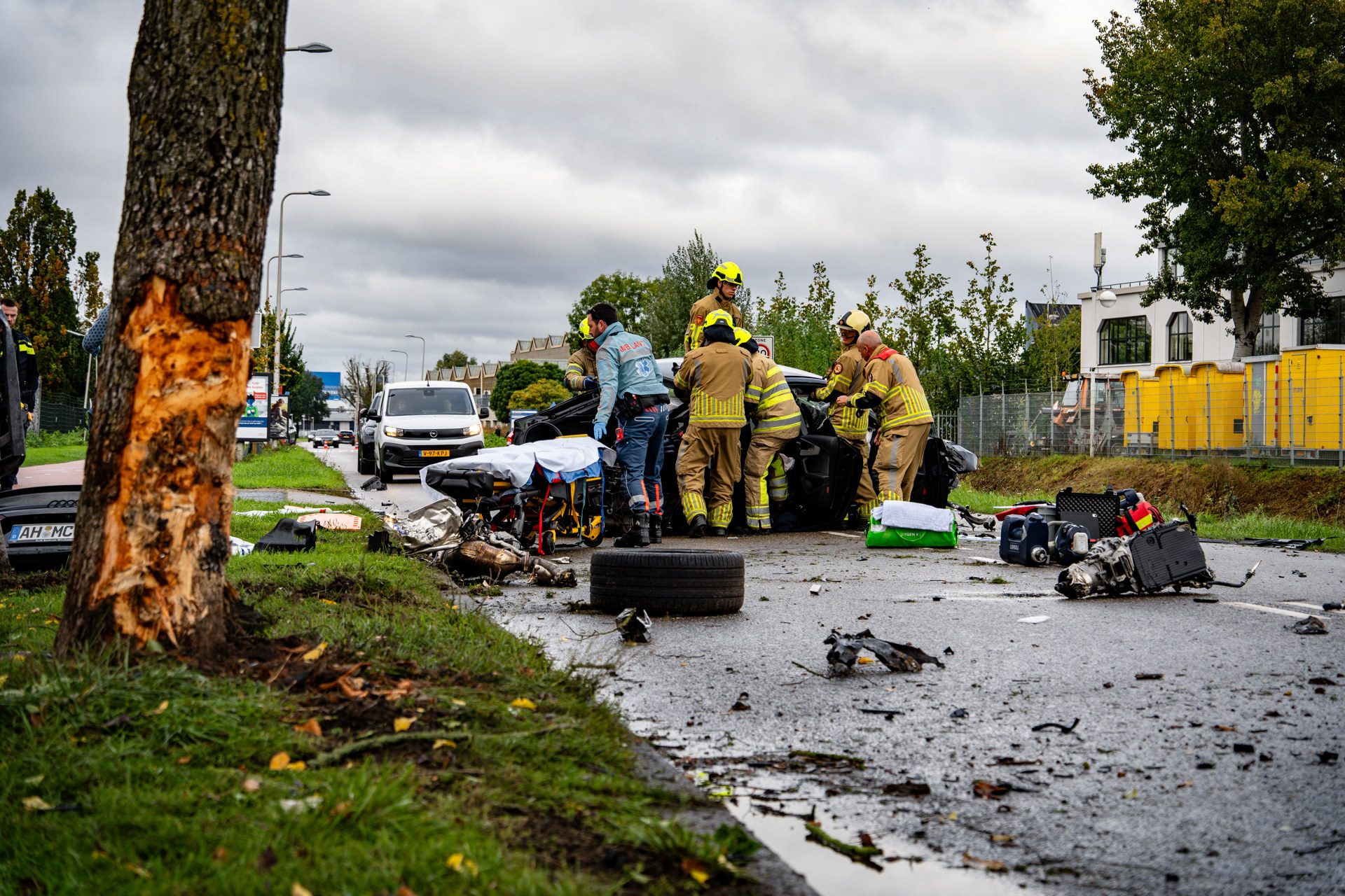 Emergency responders in yellow helmets work around a damaged car after a roadway crash, with scattered debris and equipment nearby on a cloudy day.