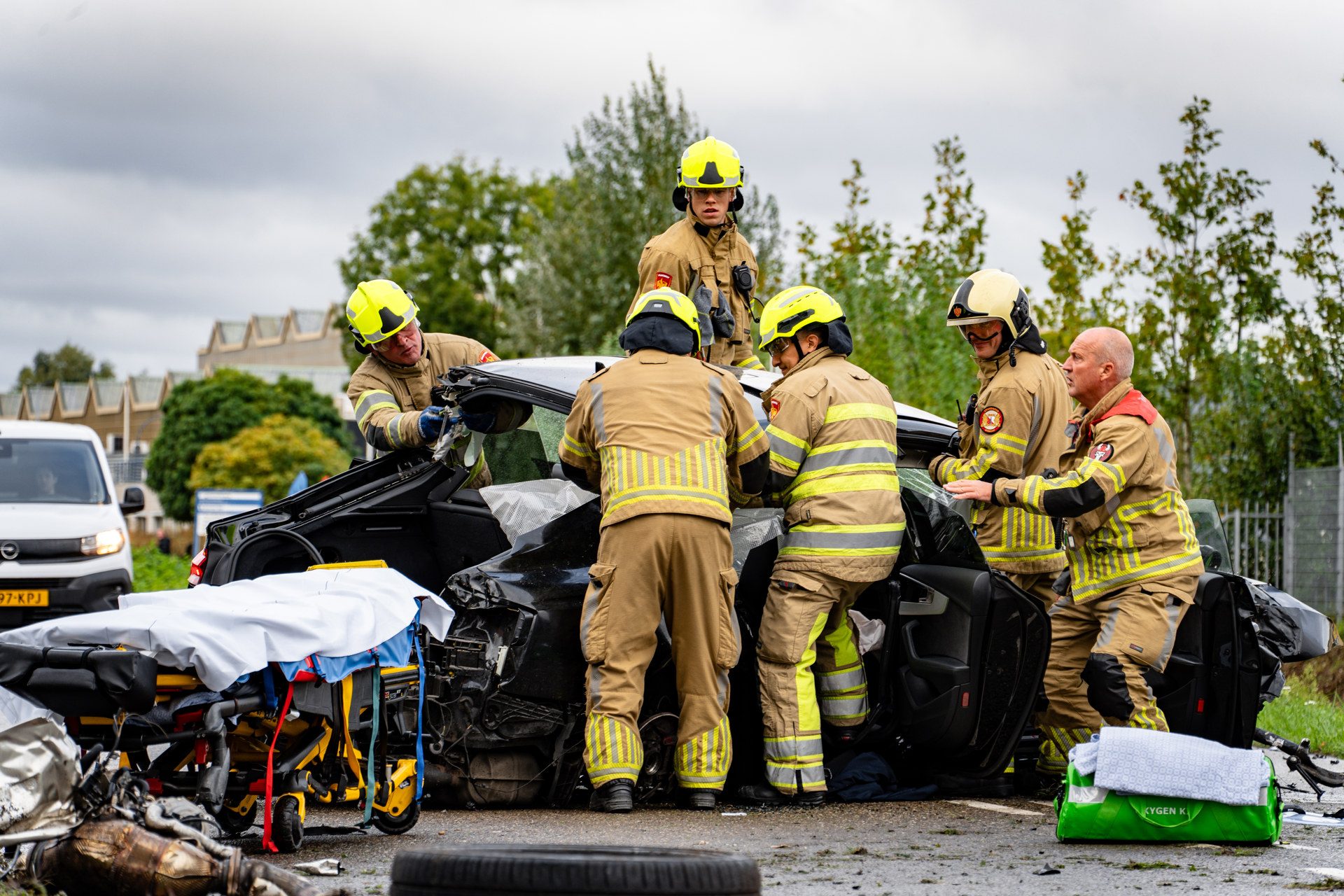 Firefighters in yellow helmets inspect a severely damaged car after a crash, with rescue gear nearby.