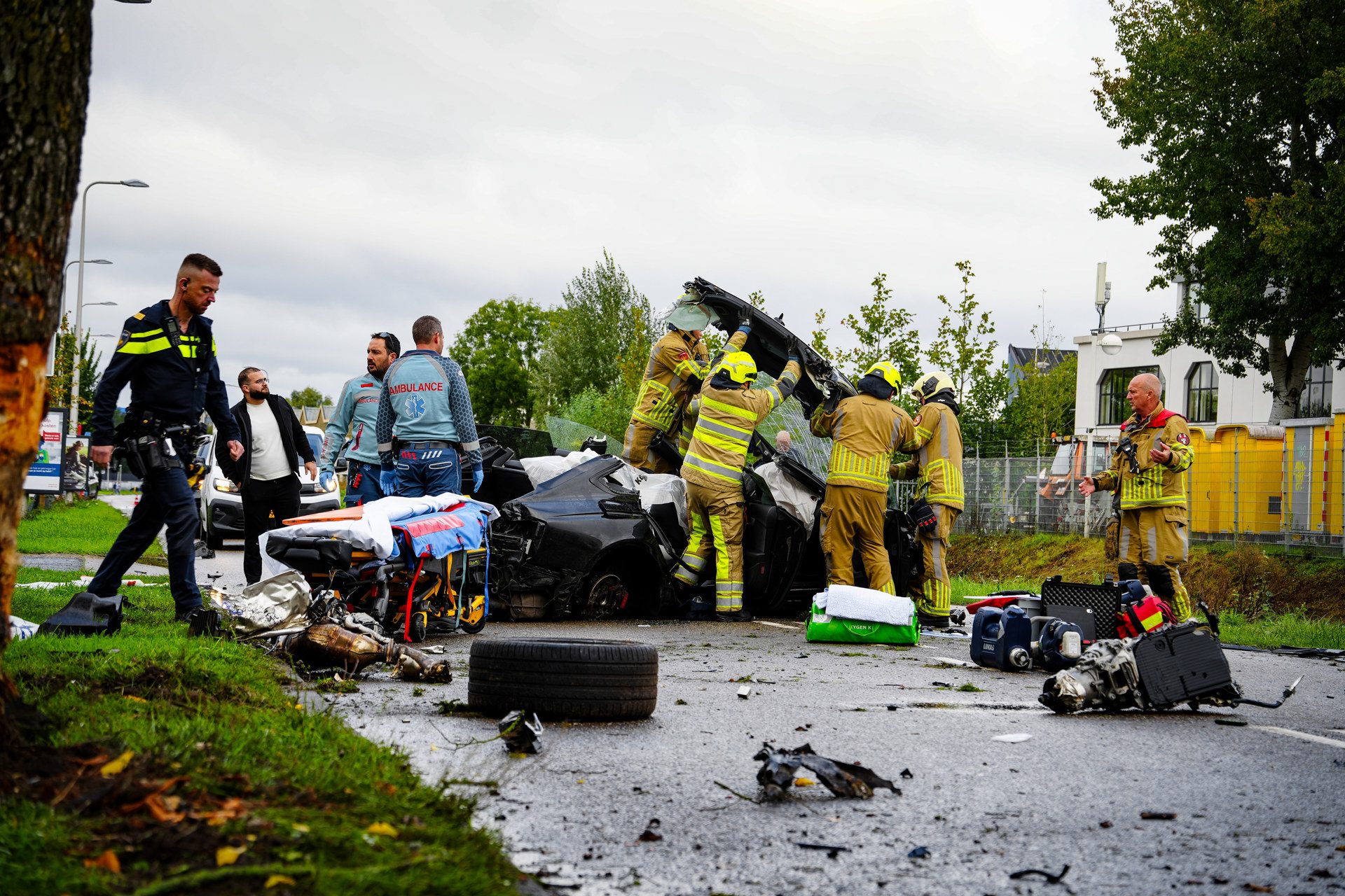 Emergency responders (firefighters and paramedics) work around a severely damaged car after a crash, with equipment and debris scattered nearby across the road.