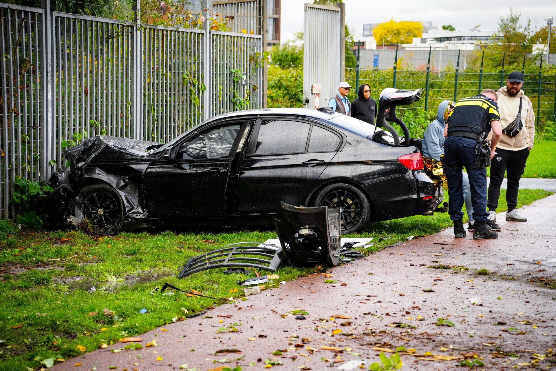 Black sedan mangled from a crash, parked on a sidewalk with debris and bystanders nearby