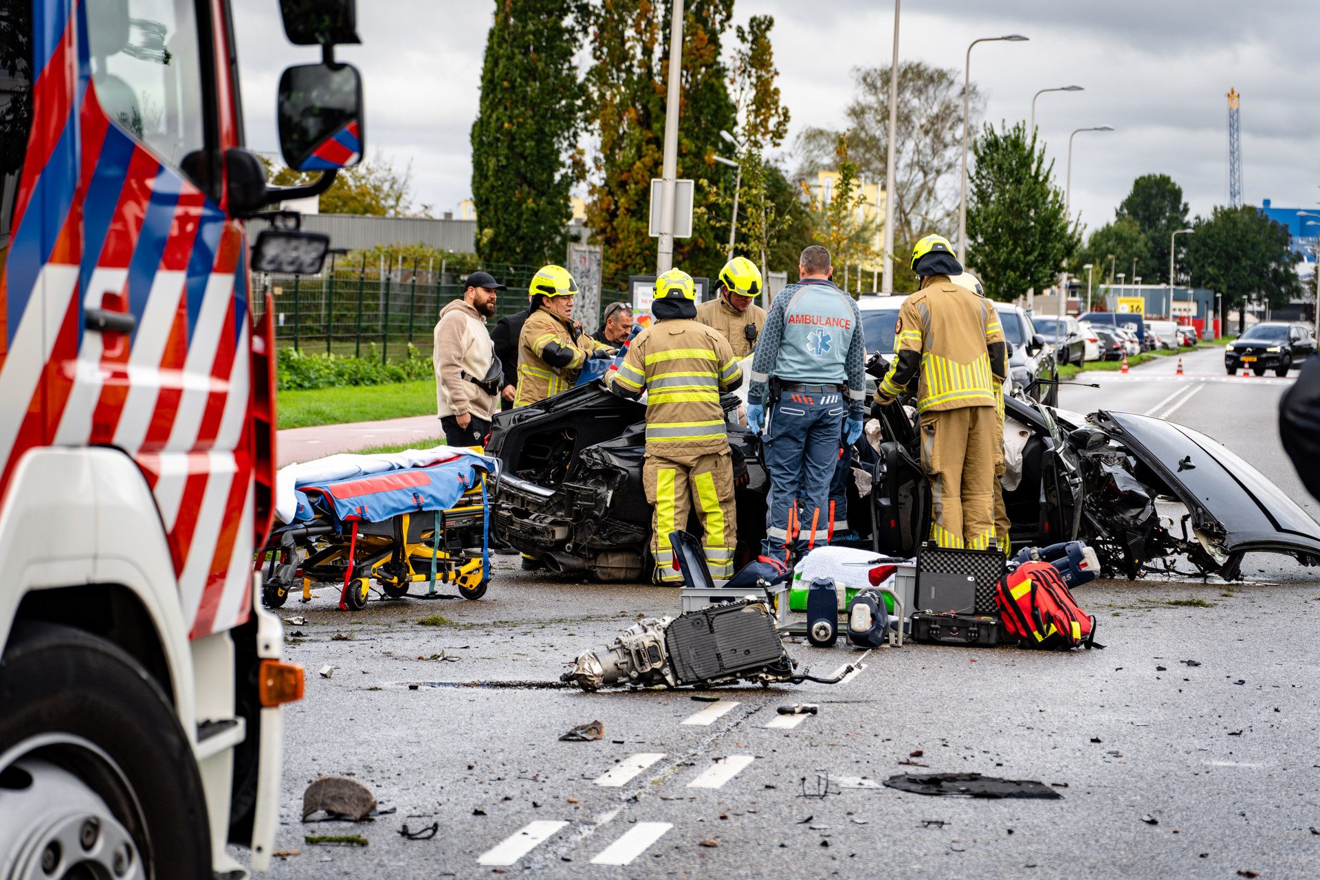 Emergency responders in yellow helmets and tan gear work around a severely damaged car on a city street, with ambulances and equipment scattered nearby.