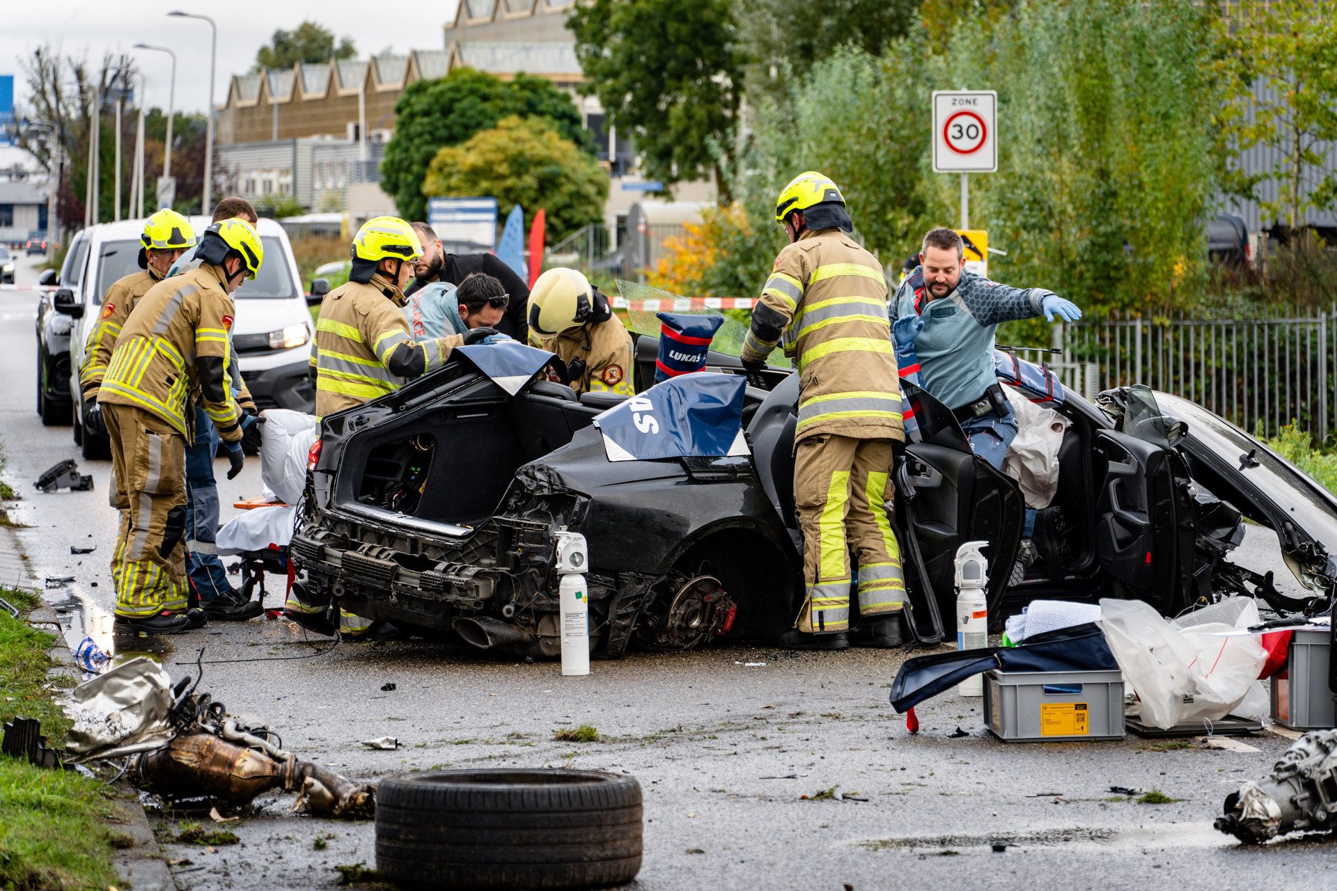 Firefighters in yellow helmets work around a severely damaged black car on a city street with debris and rescue gear nearby.