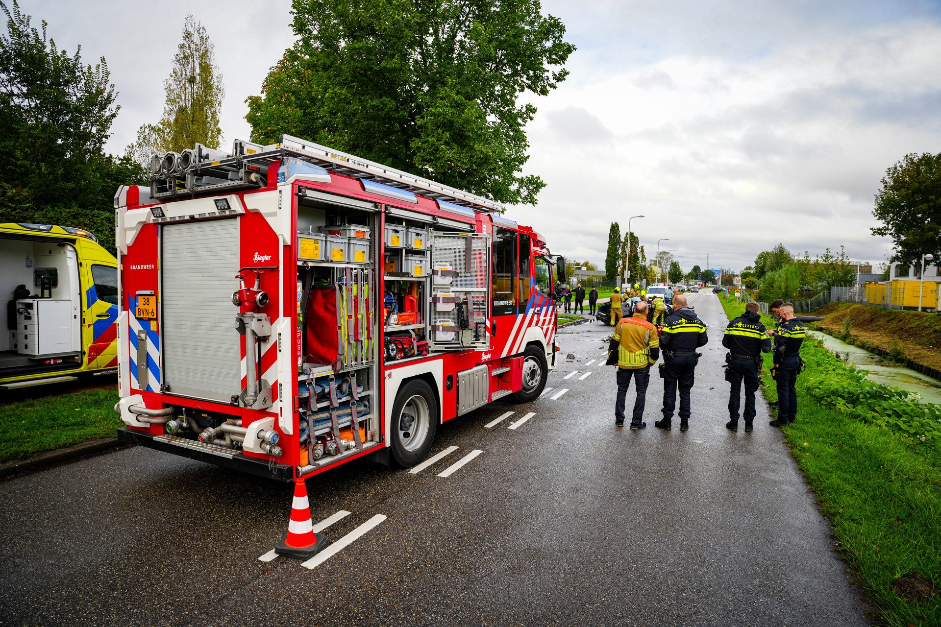 Red fire engine with open compartments parked on a wet road; firefighters in reflective gear stand nearby as an ambulance is parked in the background on a cloudy day.