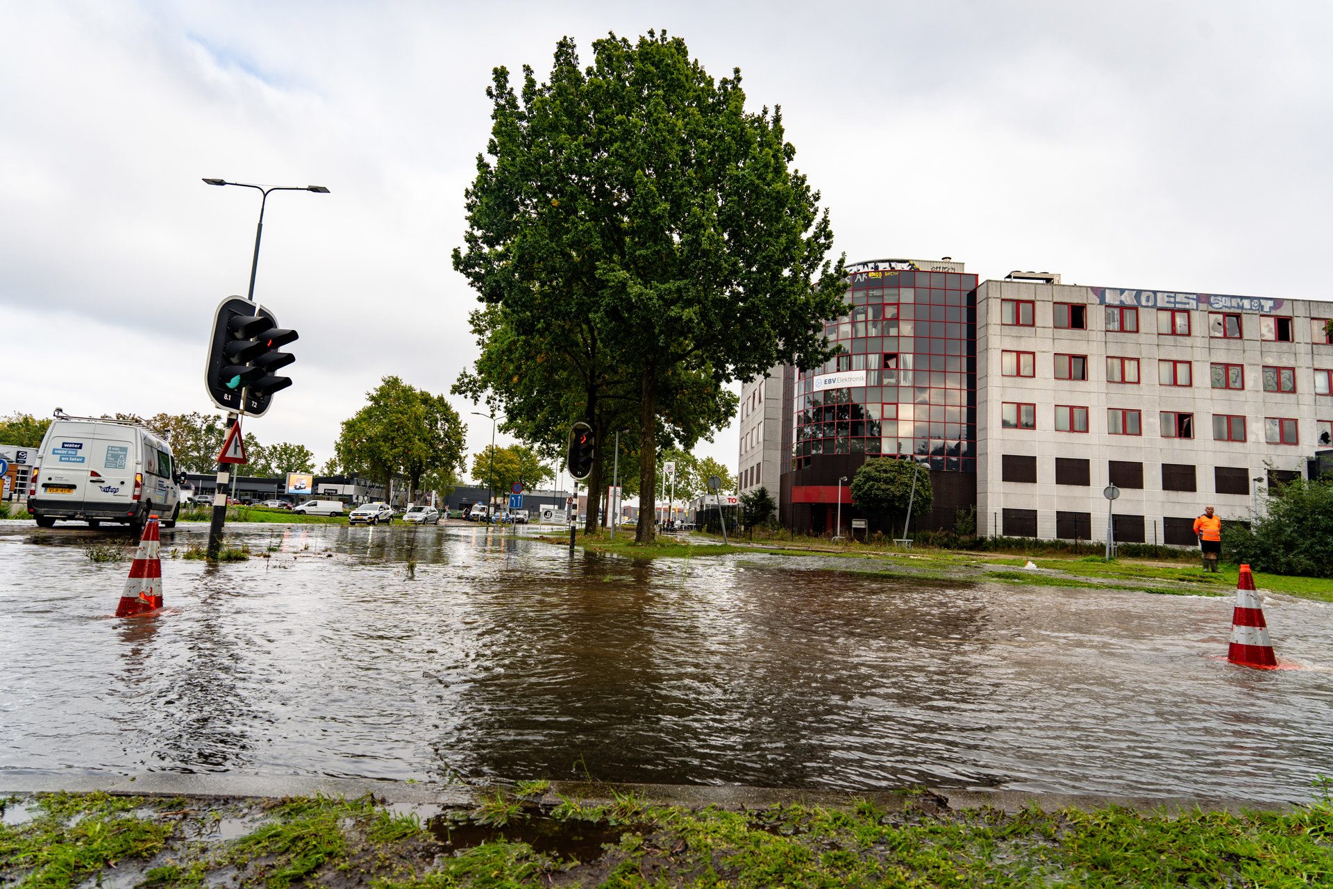 Flooded city street with water covering the road, traffic light, parked vans, and a large tree beside a modern building under cloudy sky