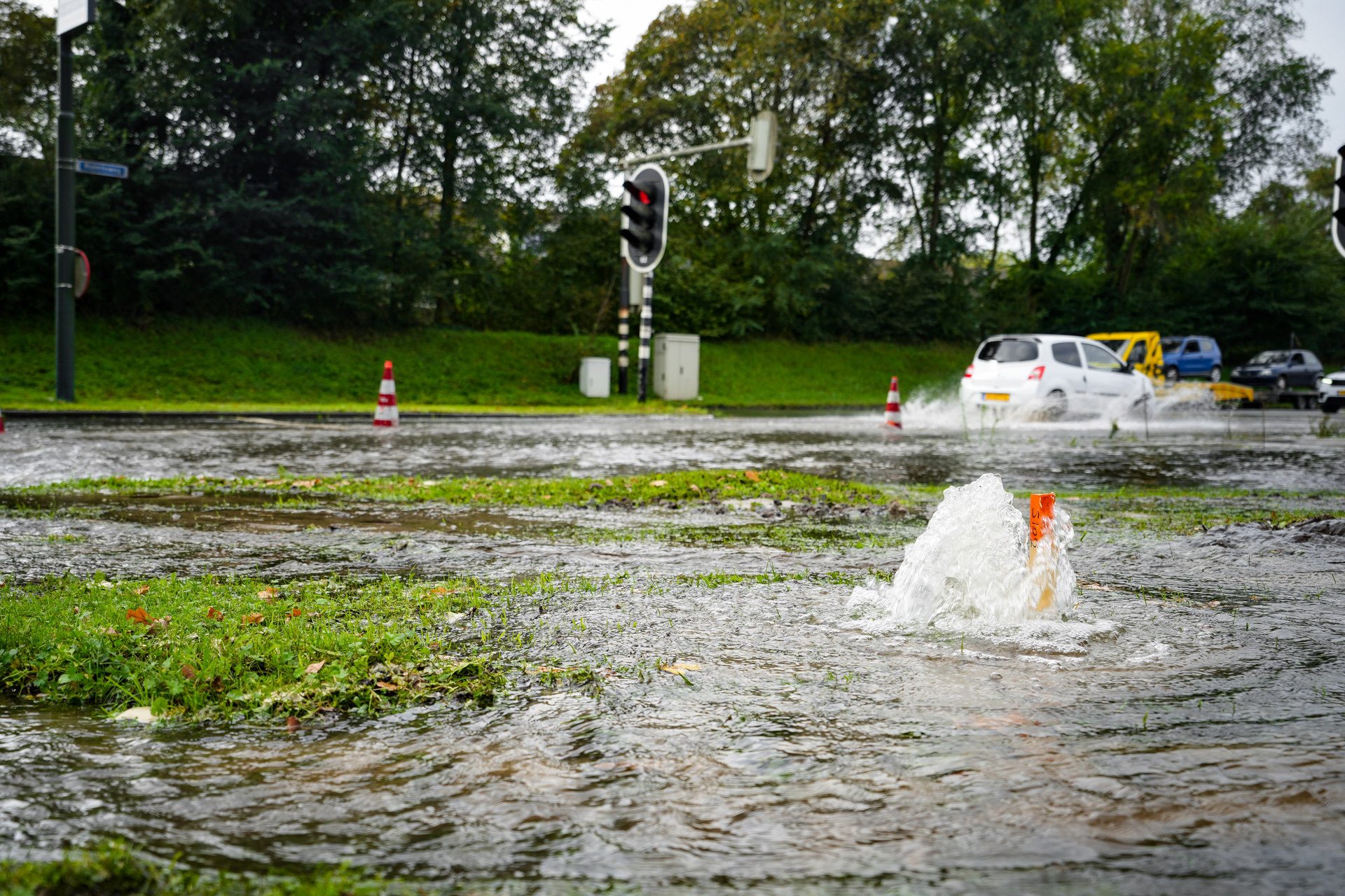 Flooded street scene with moving white car splashing water, orange cones, and a red traffic light in the background.