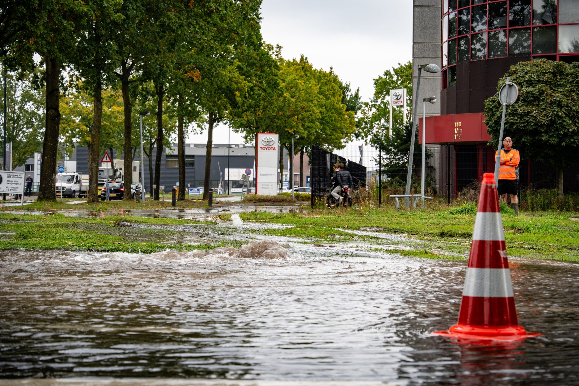 Flooded urban street beside a Toyota dealership with a man in an orange safety vest standing near a striped orange cone and muddy water in the foreground.
