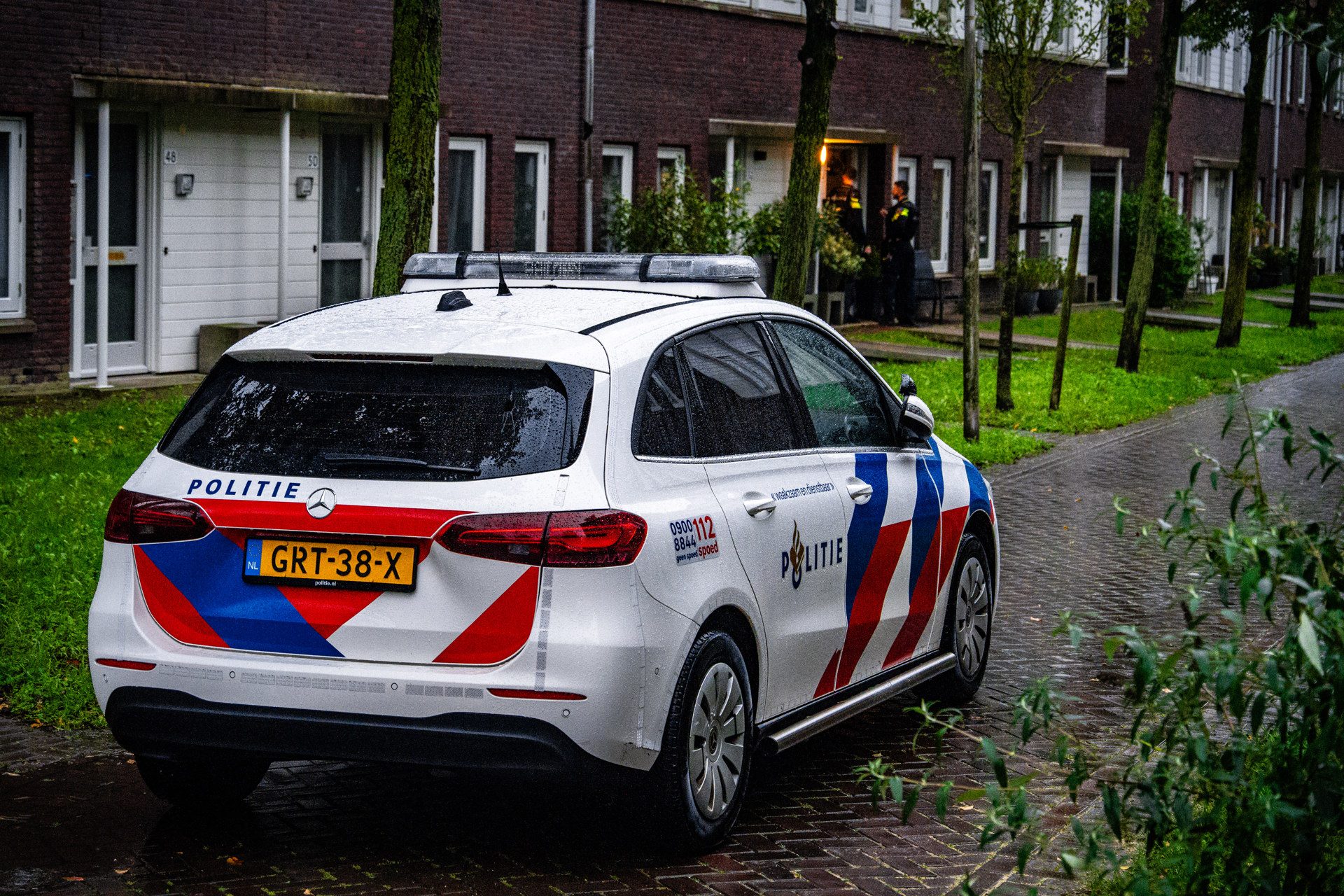 Rear three-quarter view of a Dutch Politi e police car with blue-red chevrons parked on a wet brick street in a residential area; two officers stand near an open door in the background.
