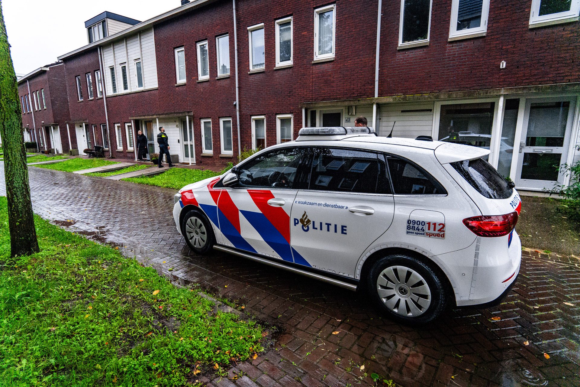 Dutch police car parked on a wet brick street in front of row houses, with officers near the entrance of a home, daytime scene.