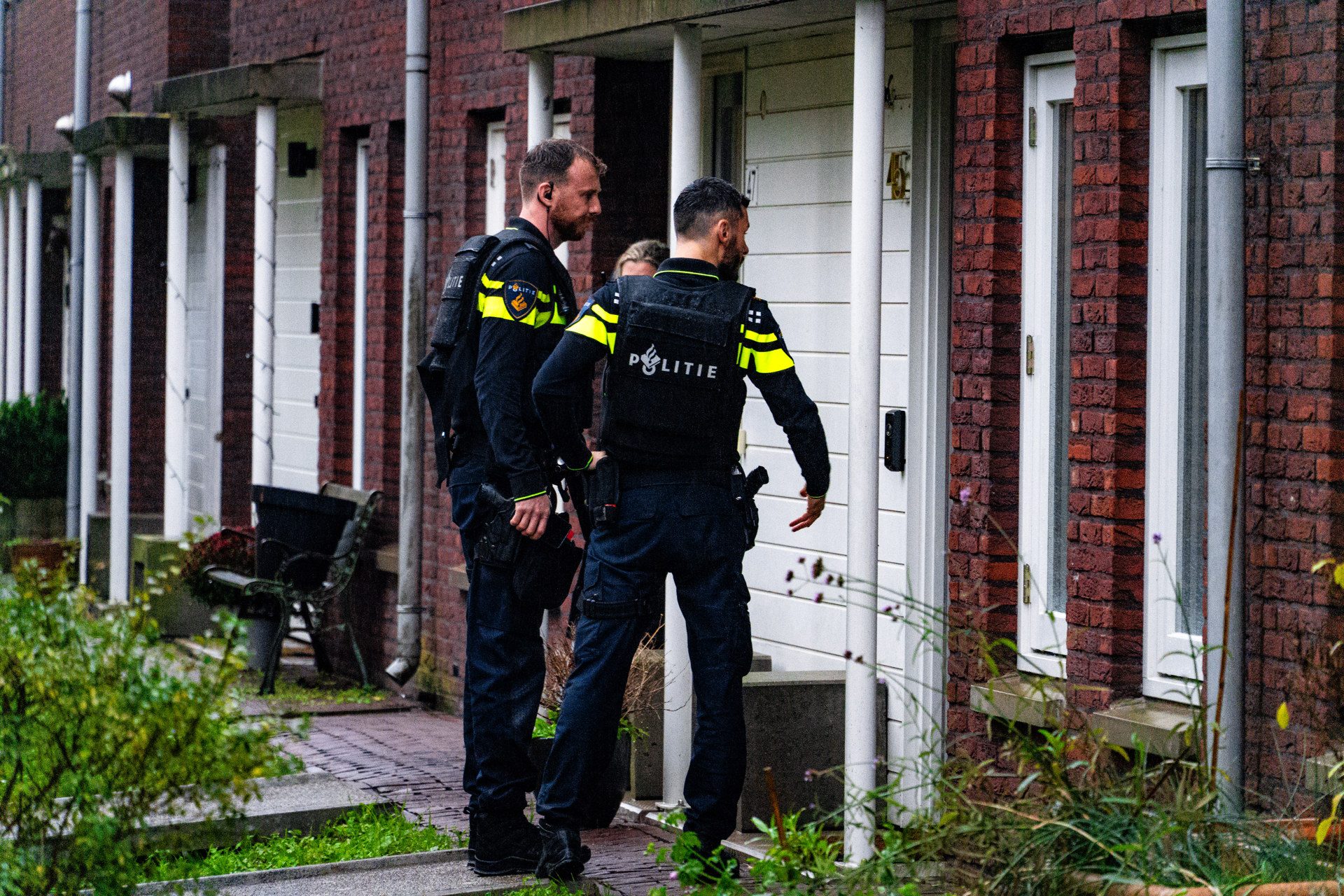 Police officers in tactical gear with neon-yellow stripes stand on a sidewalk outside a row of brick townhouses, near white doors.