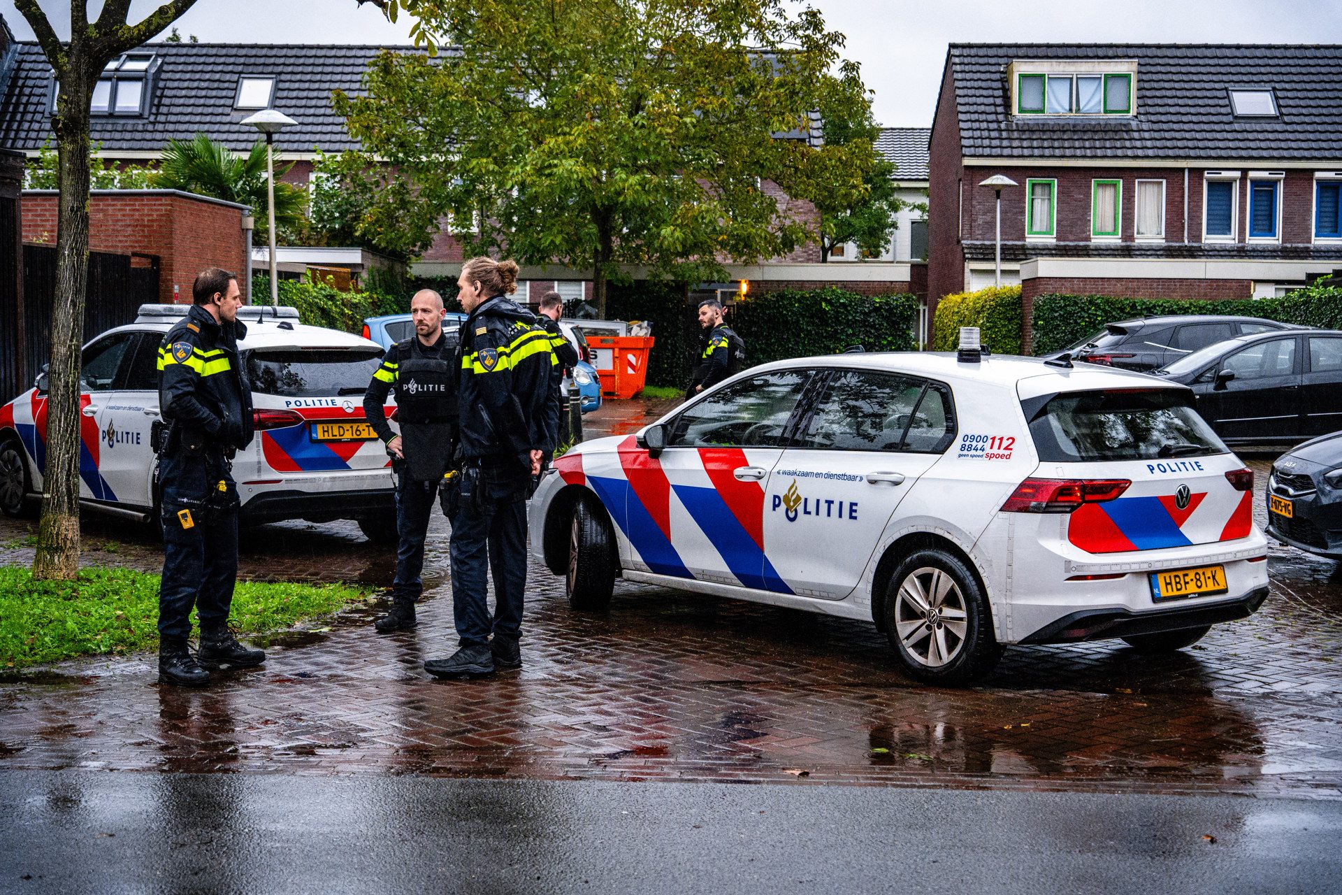 Group of Dutch police officers in dark uniforms standing and talking beside marked police cars on a wet residential street after rain.