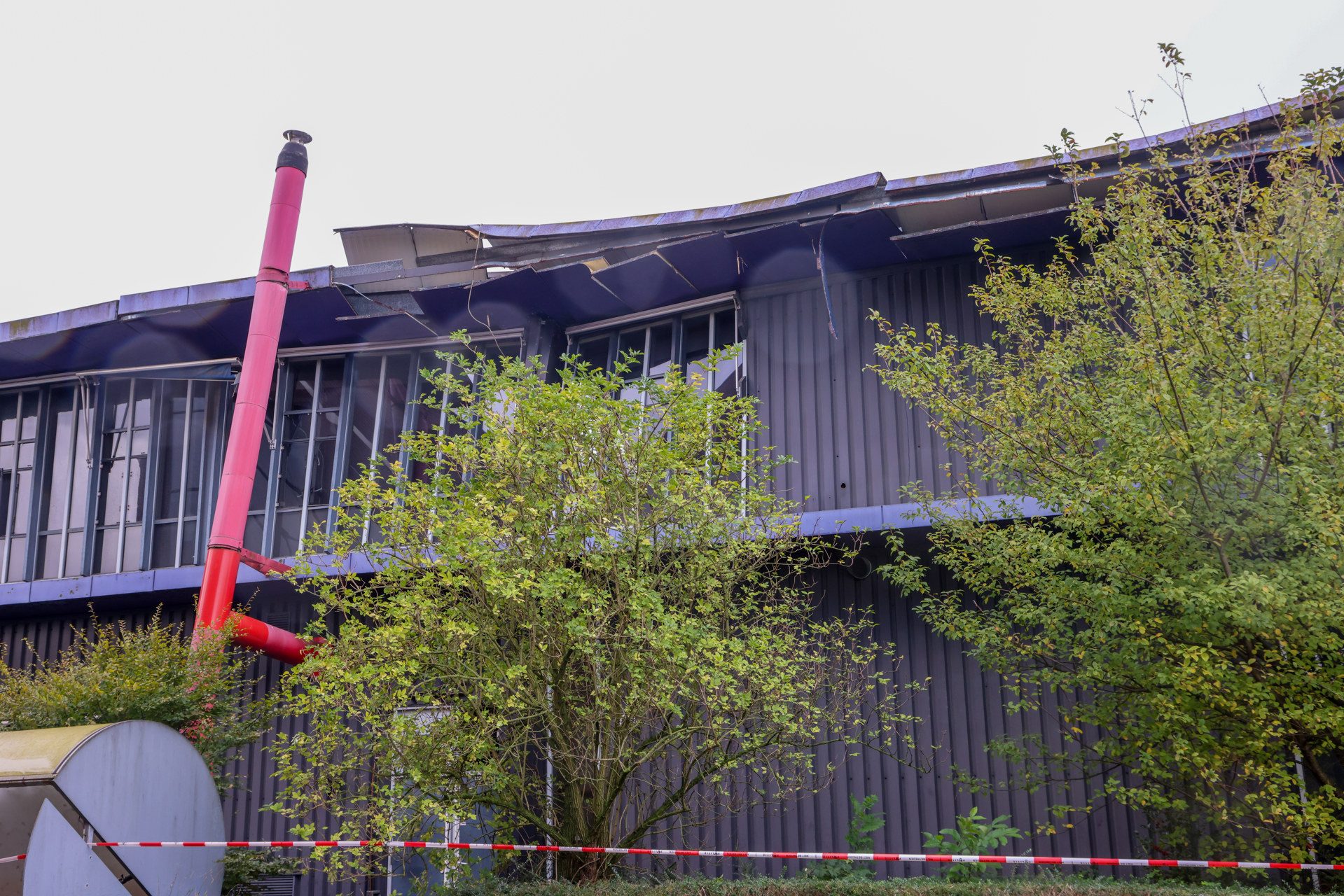 Industrial building with a severely damaged roof and exposed metal, a pink emergency pipe, and overgrown trees in front; red caution tape on the ground.
