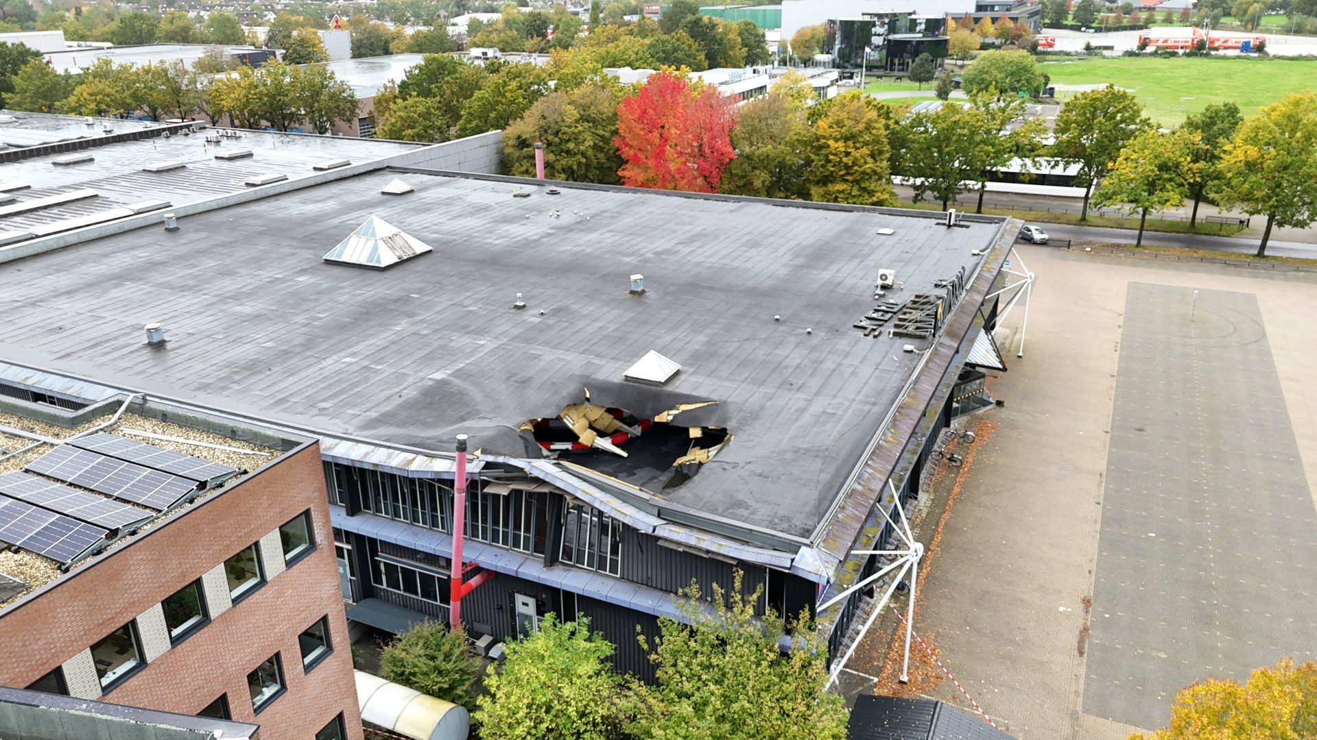 Aerial view of a flat roof with a large damaged area and collapsed material near the edge of a building, with trees and a parking area nearby.