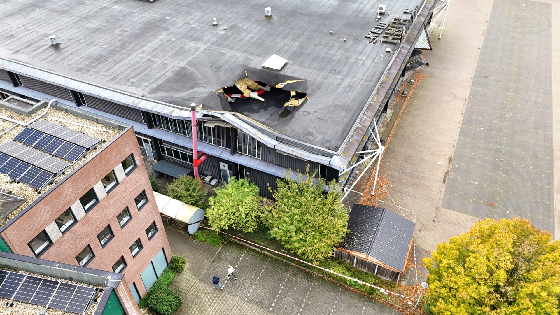 Drone view of a flat-roofed building with a large collapsed hole in the roof and exposed debris, surrounded by safety tape and nearby structures with solar panels on another roof.