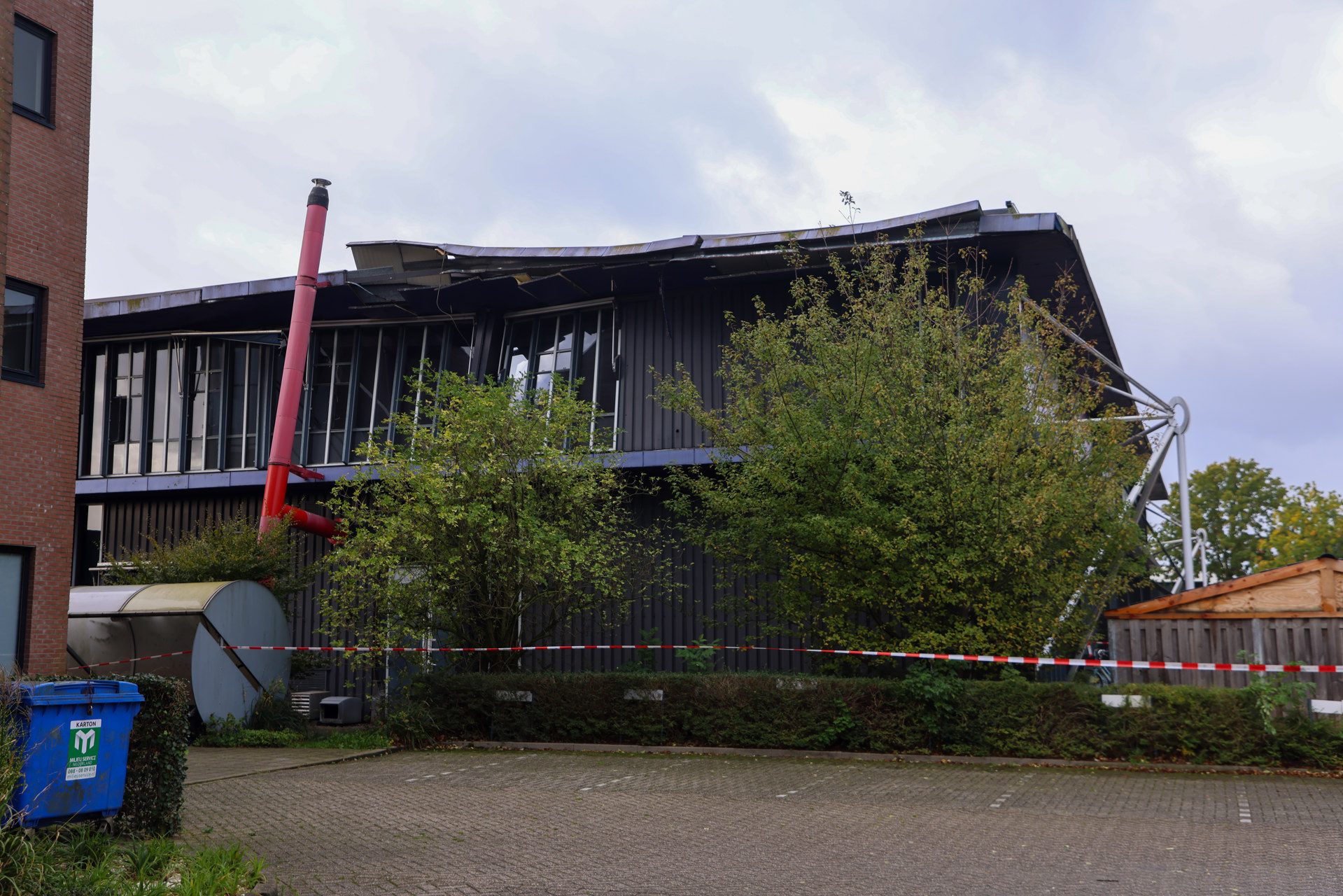 Two-story building with a severely damaged roof and broken windows, surrounded by trees and safety tape in the foreground.