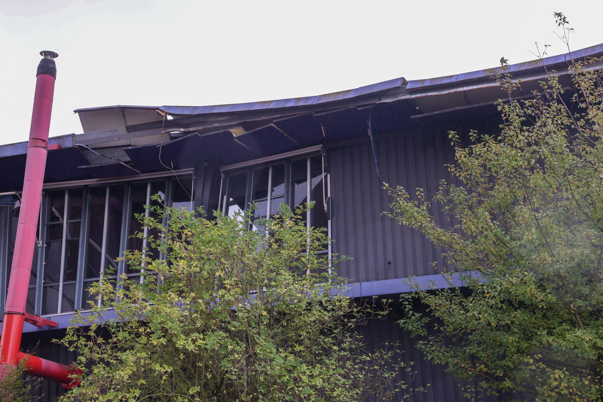 Derelict industrial building with a collapsed roof, a pink vertical pipe, and green trees in front.