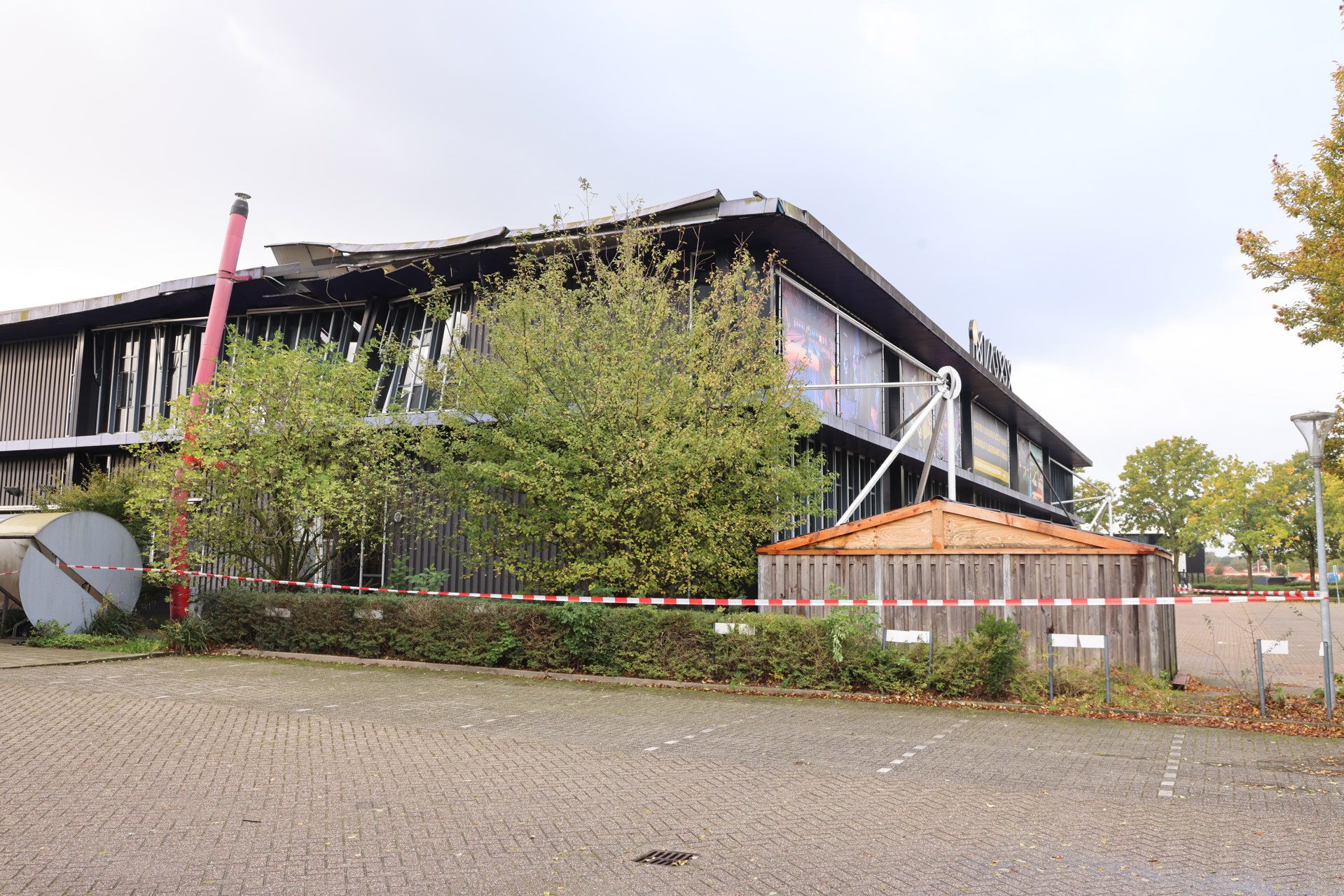 A damaged two-story building with a bent roof, red construction pipe, and red-and-white barrier tape in a parking lot, with trees in front.