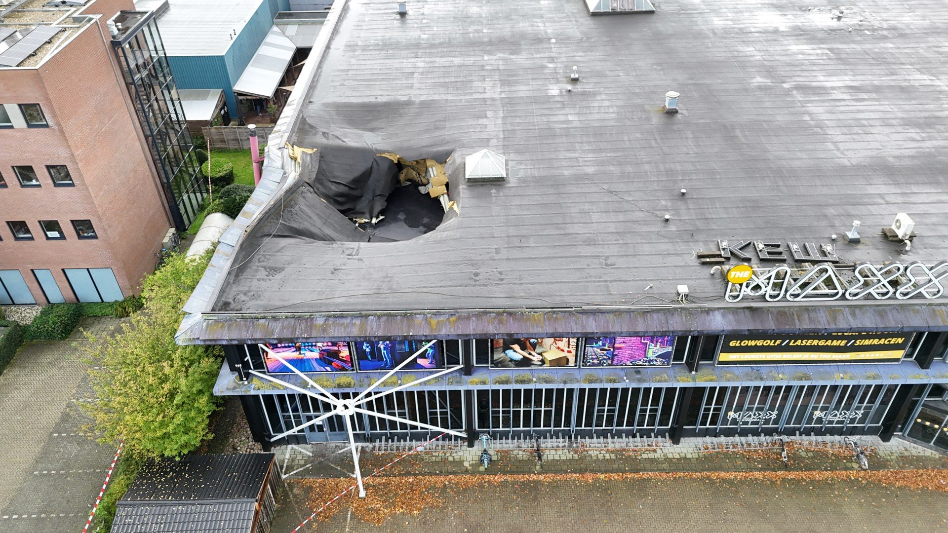 Aerial view of a flat commercial rooftop showing a large collapsed area with exposed material and a puddle of water near the center, beside a brick building with glass walls.